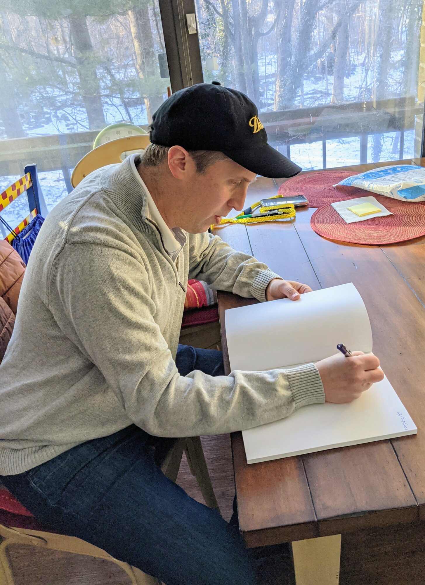 Person sitting at a table, writing in a large open notebook, wearing a black cap and light-colored sweater.