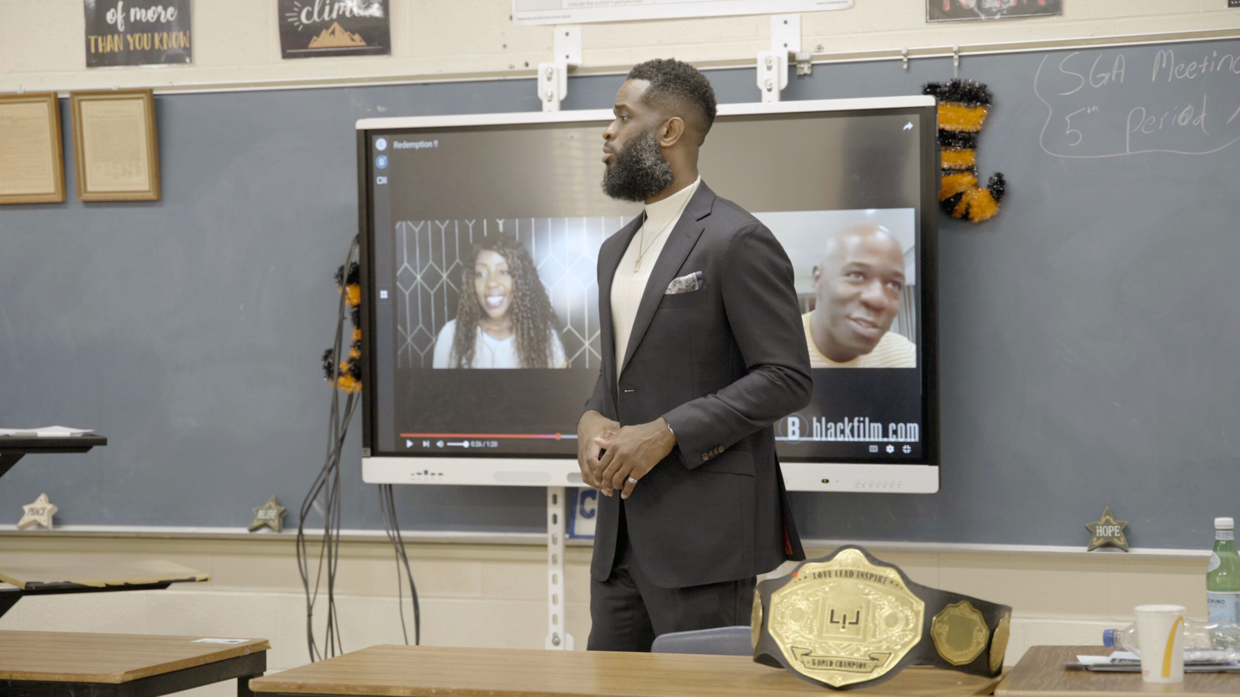 Man in suit standing in classroom with digital screen showing two people, a blackboard, and a championship belt on desk.