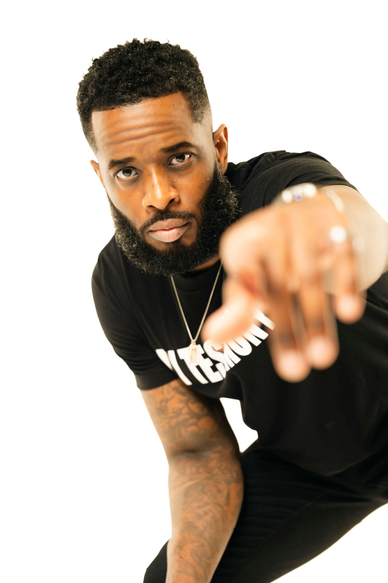 Man with short curly hair and beard pointing towards the camera, wearing a black T-shirt, against a white background.