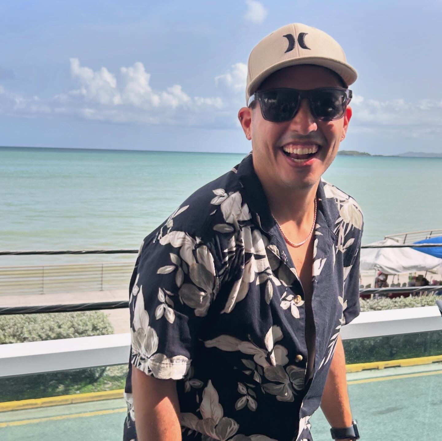 Man wearing sunglasses, a beige cap, and a floral shirt smiling outdoors near water and a railing.