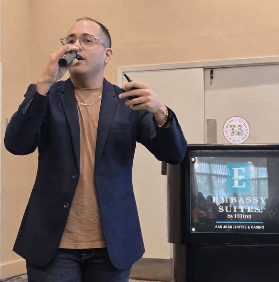 Man speaking into microphone, holding phone, wearing glasses, blazer, and tan shirt, standing indoors near a sign and screen.