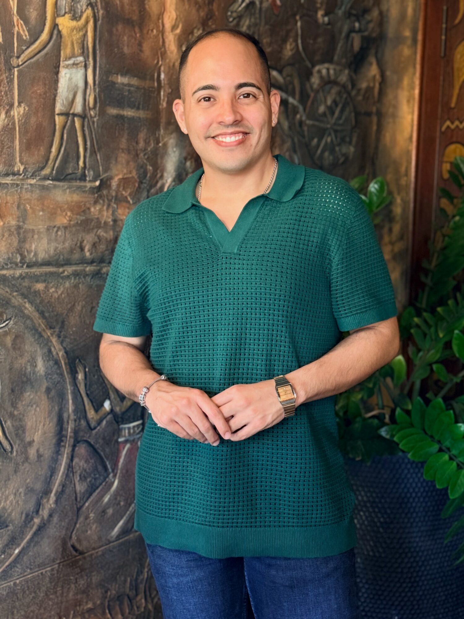 Smiling man in green polo shirt and jeans standing indoors with a mural and plants behind him.