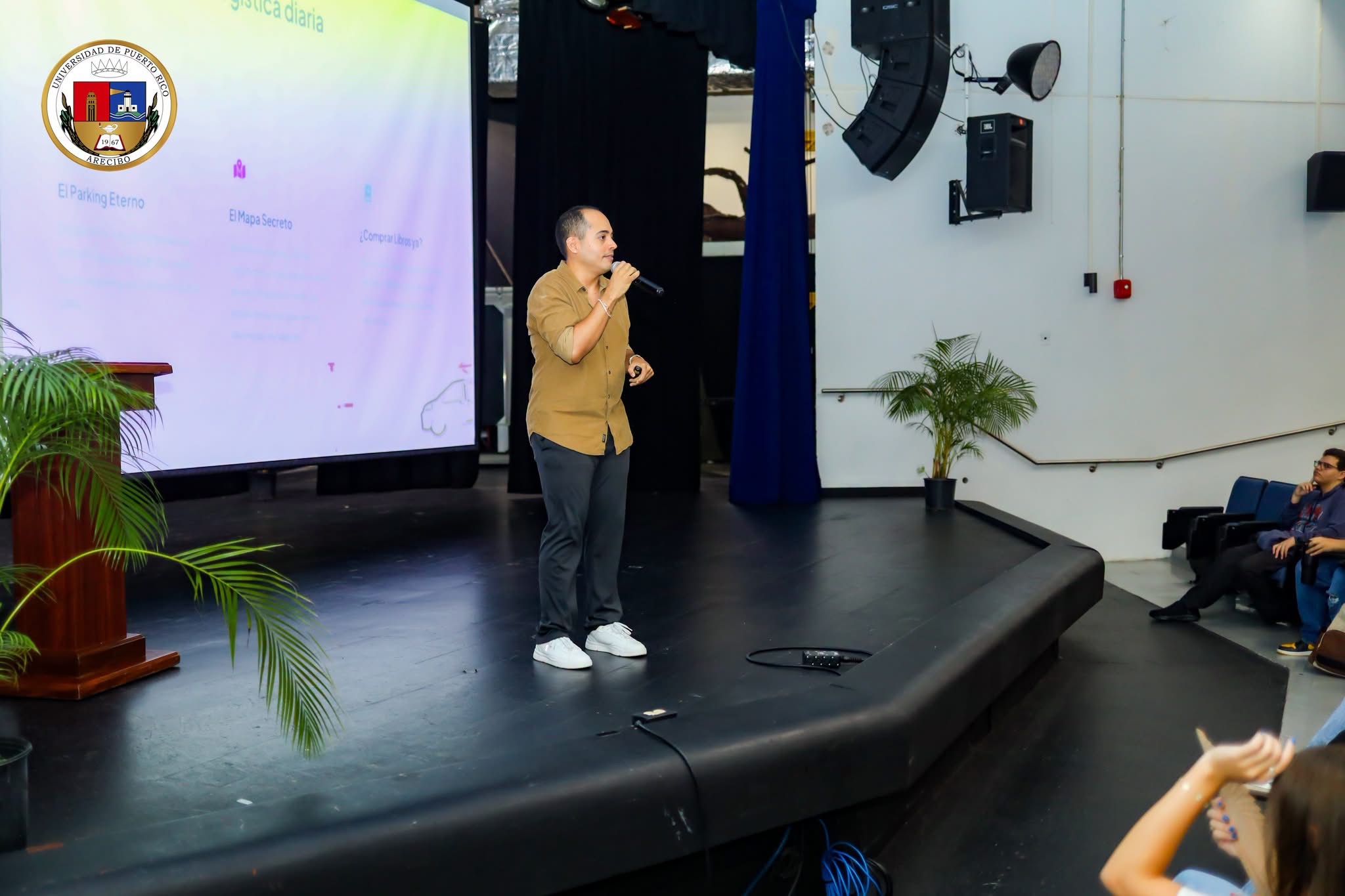 Person speaking on stage with a microphone in a conference room, audience seated, large screen behind, plants on stage, and speakers mounted on wall.