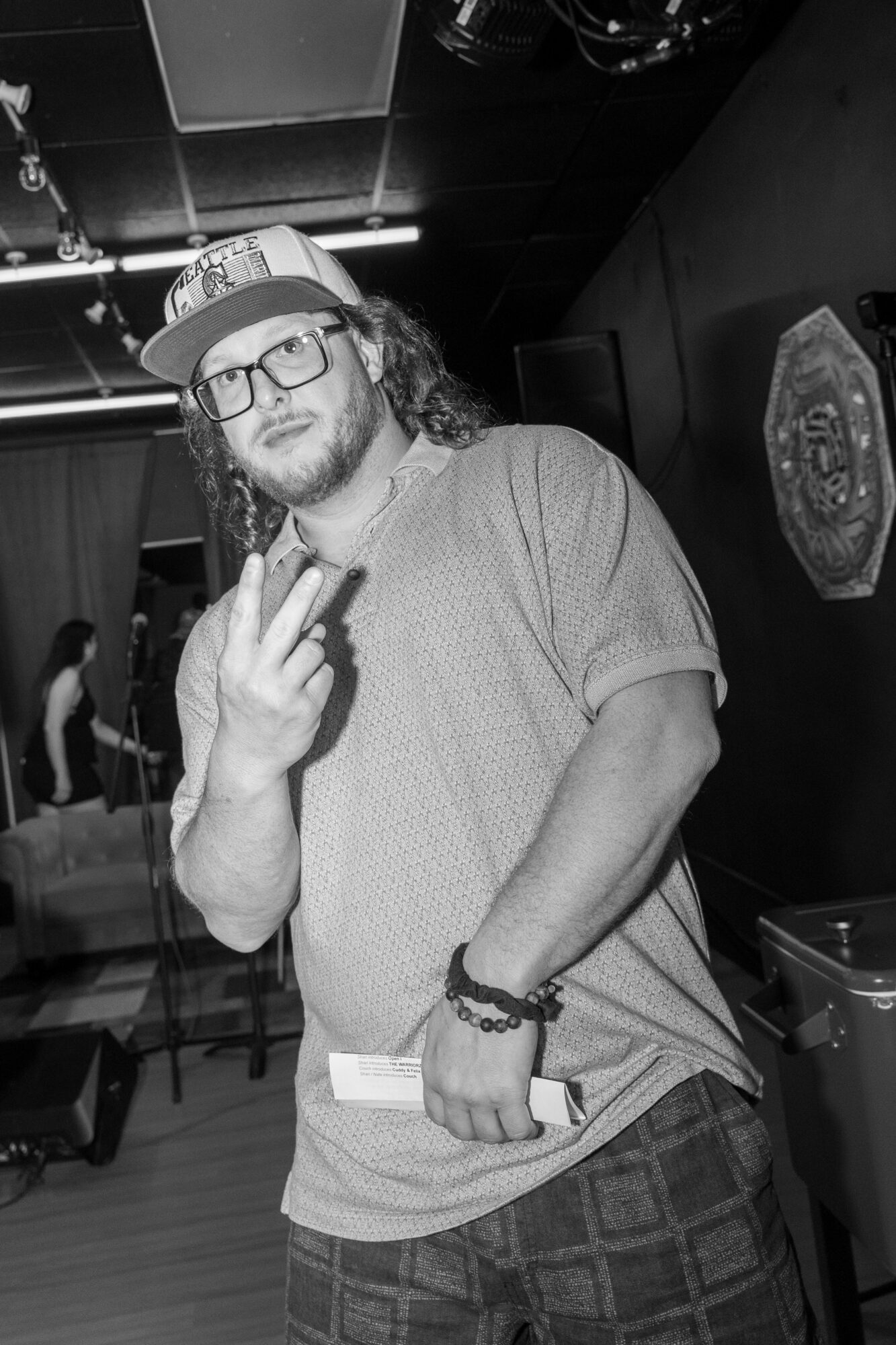 Man with glasses and a cap making a peace sign, standing indoors with a dark background.