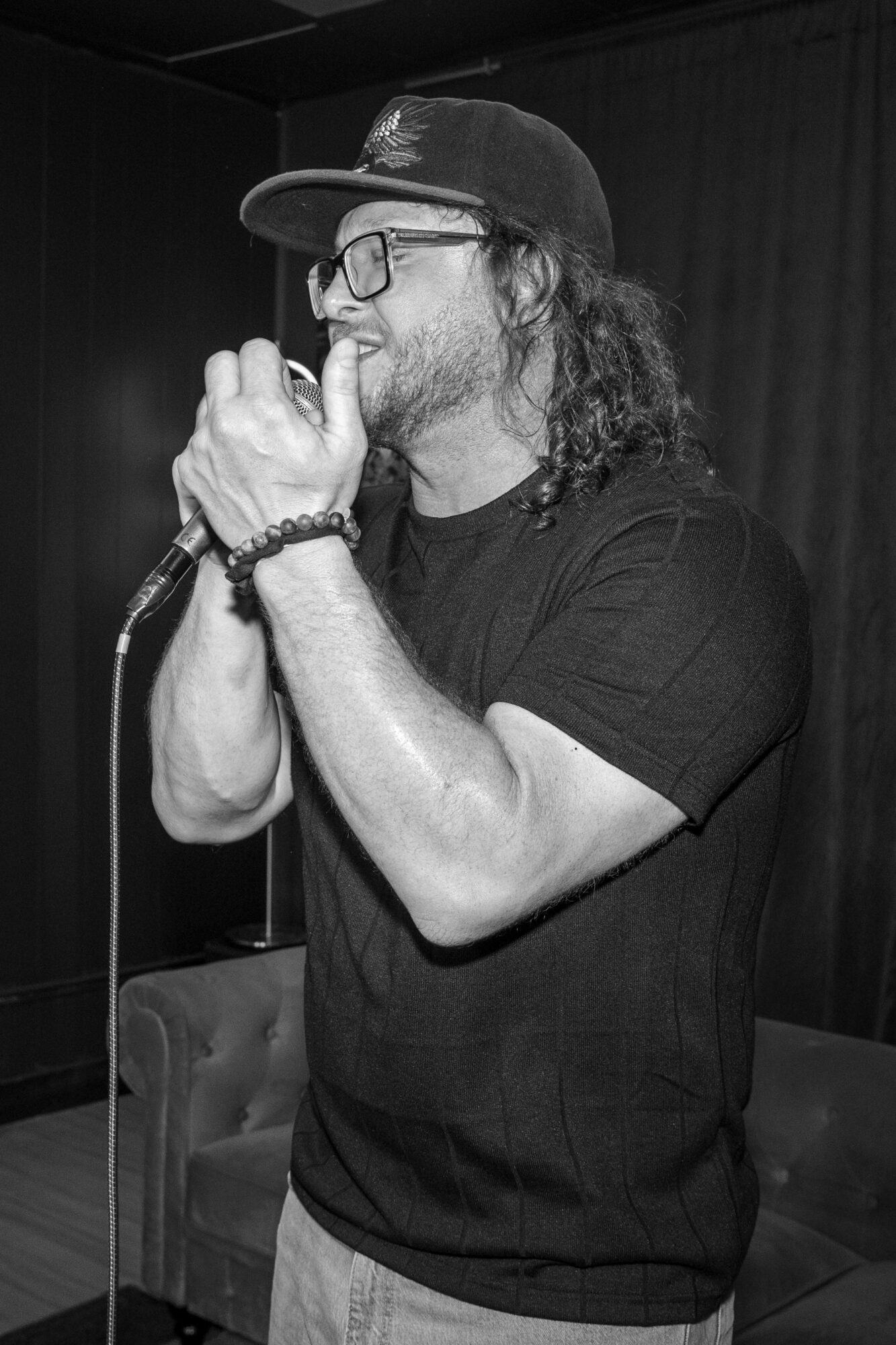 Man with glasses and a cap holding a microphone, smiling, in a room with dark background and furniture.