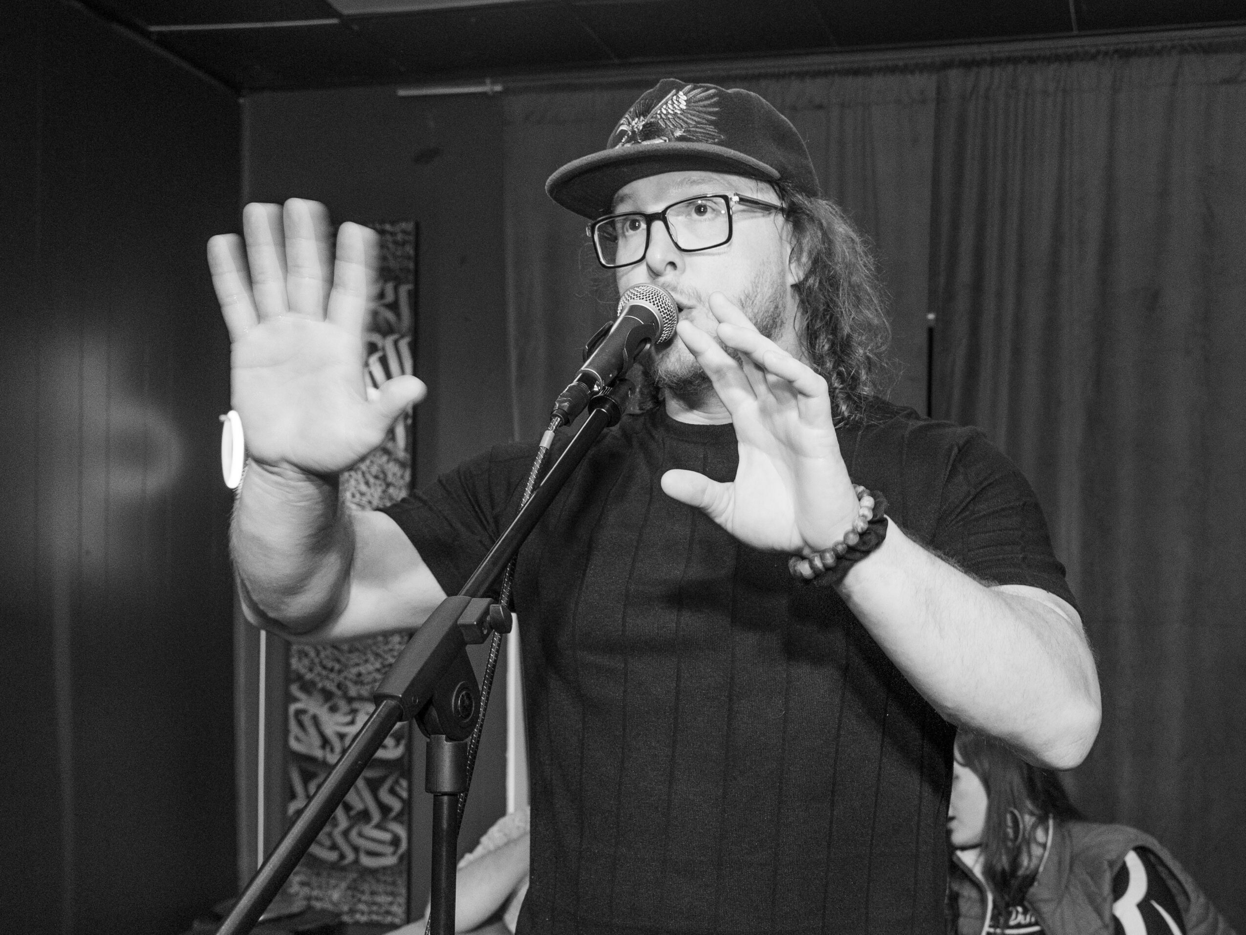 Man with glasses and a hat speaking into a microphone, gesturing with hands, in a performance setting.