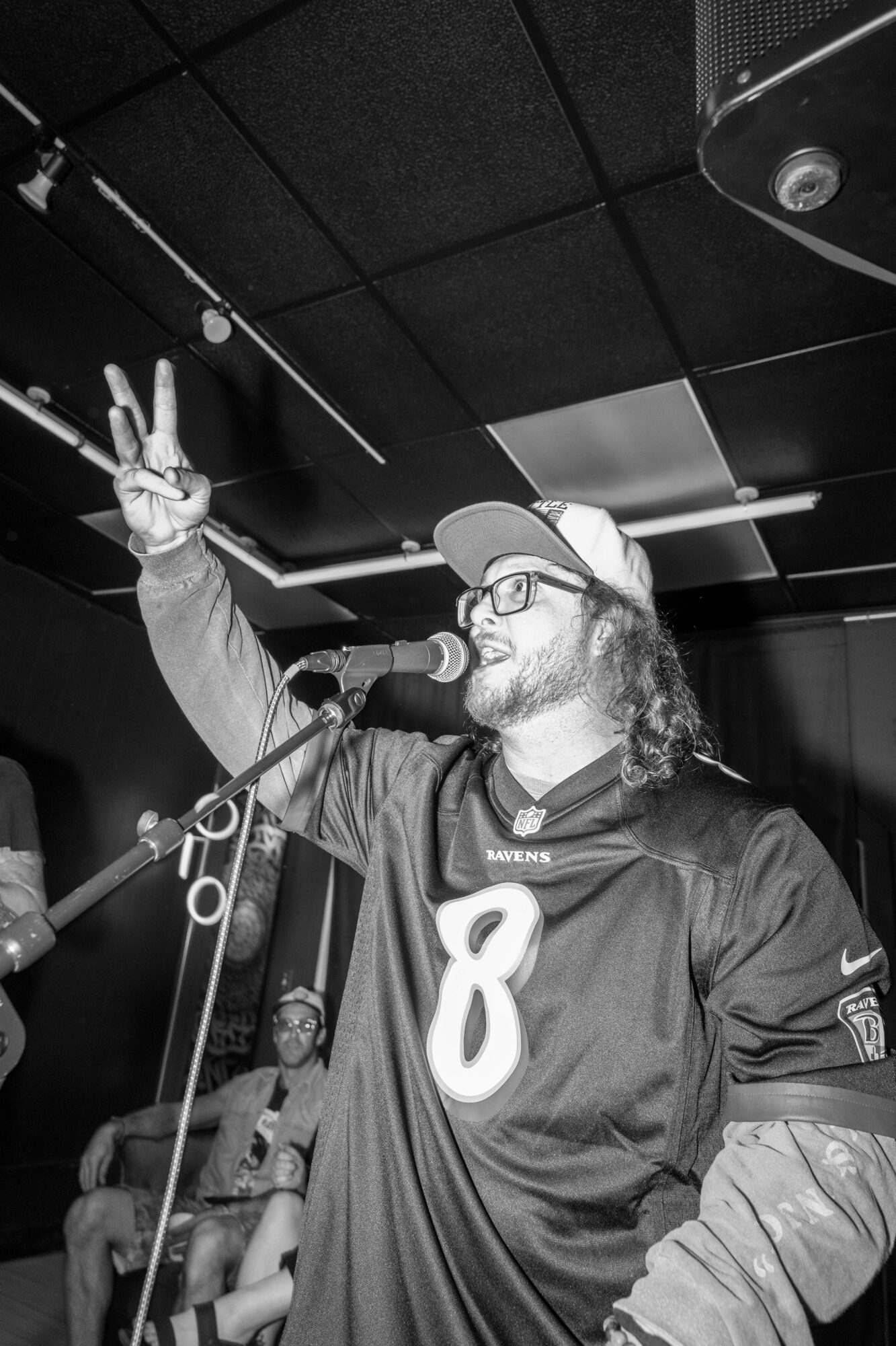 Man with glasses and a beard wearing a sports jersey and cap, speaking into a microphone, raising his hand with a peace sign, in a dark room