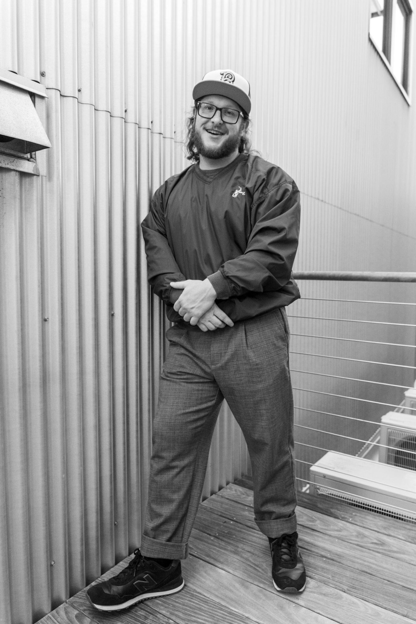 Person with glasses and a cap smiling, standing outdoors against a corrugated metal wall, wearing a jacket and pants.