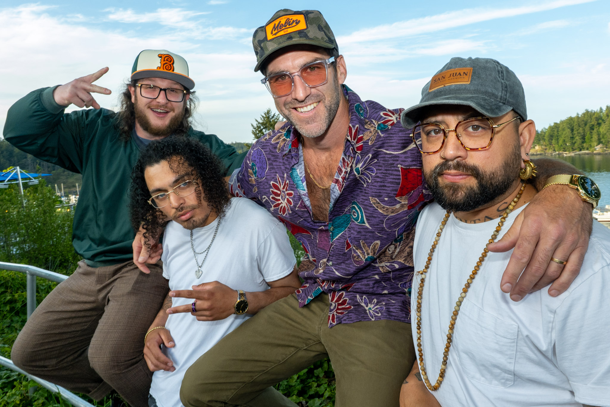 Four people outdoors smiling, with a blue sky and trees in the background. One person makes a peace sign, others pose happily.