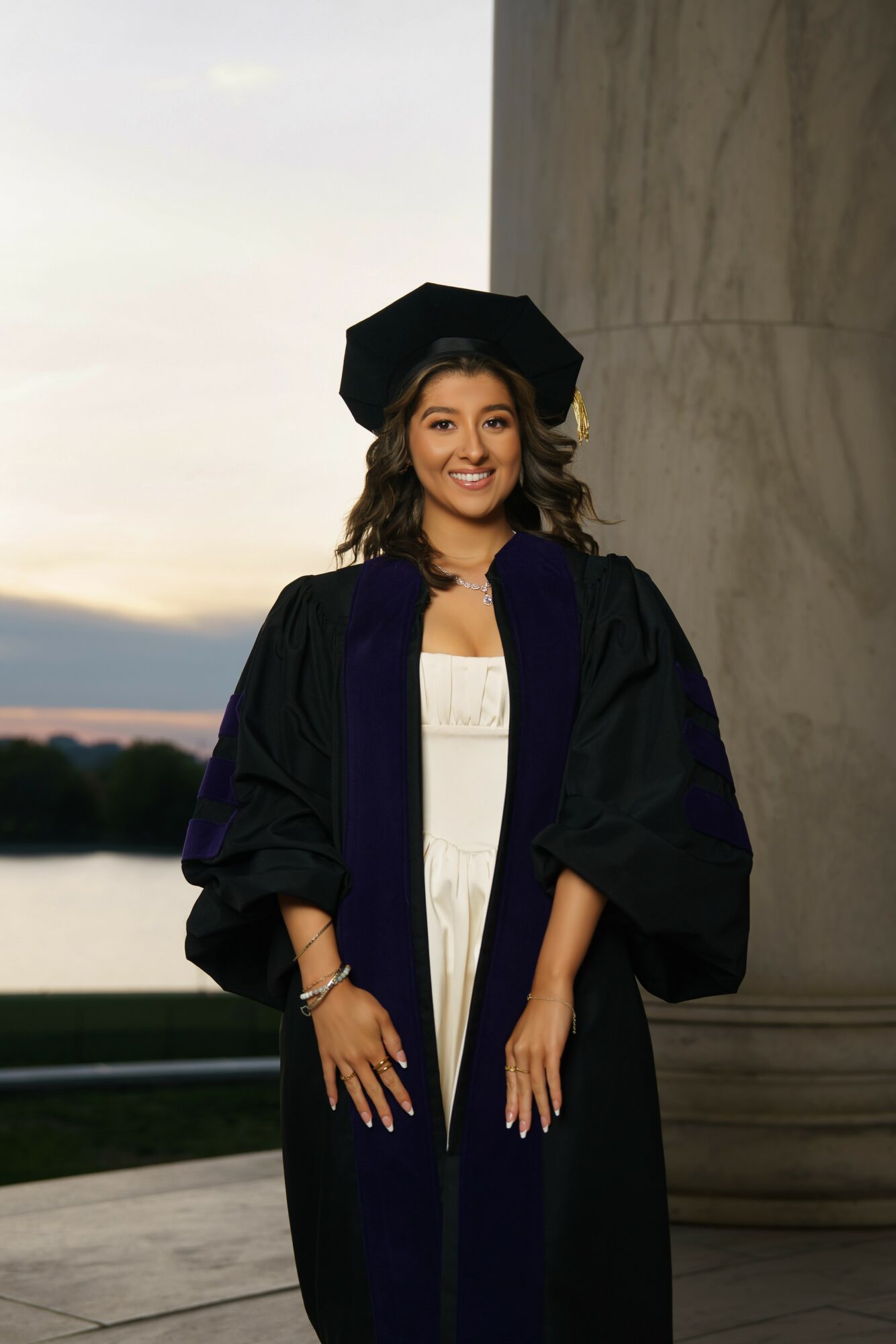 Young woman in graduation gown and cap smiling outdoors near a stone structure at sunset.