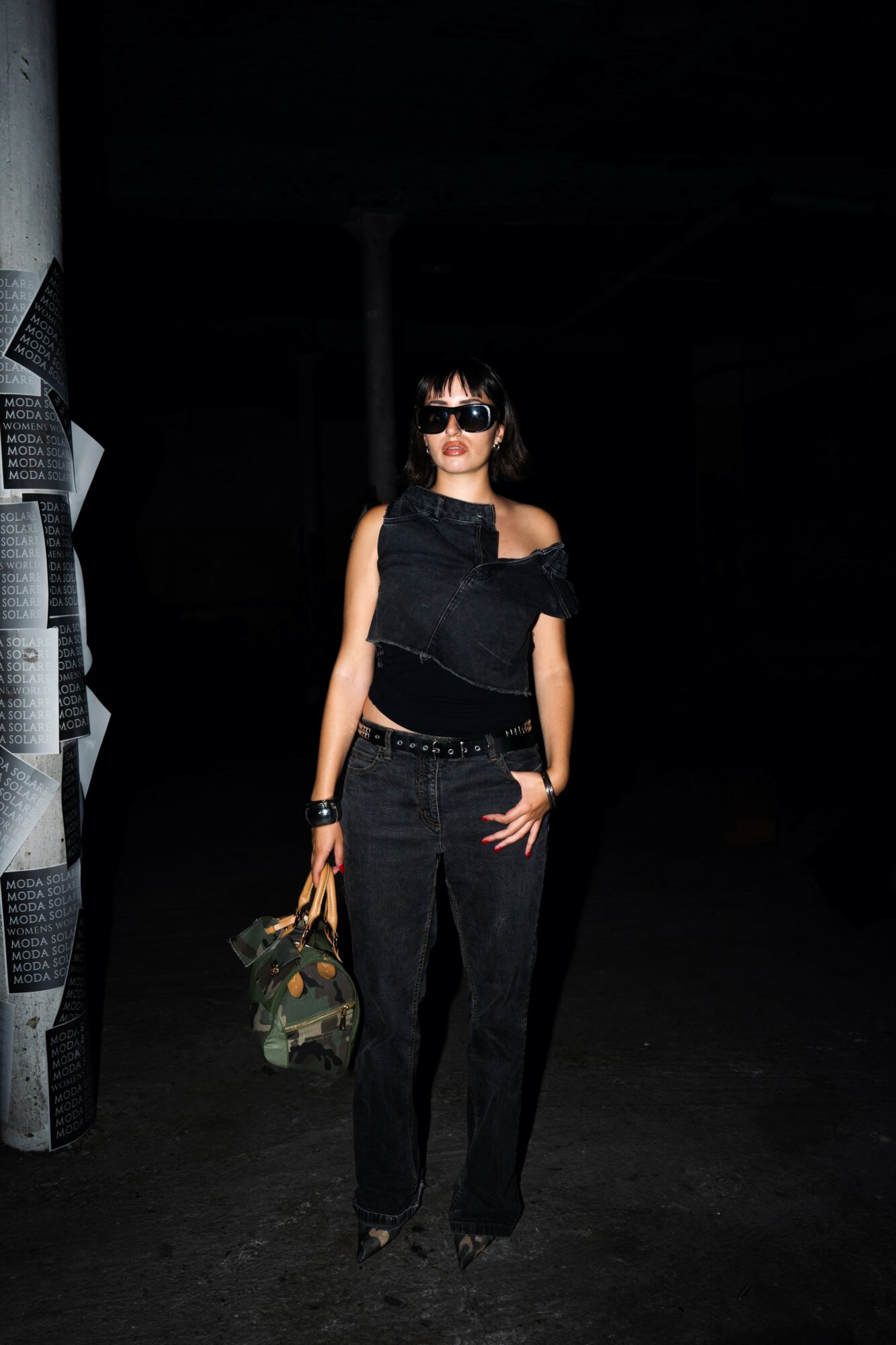 Woman with dark hair and sunglasses standing in a dark space, wearing black clothing and holding a camouflage bag.