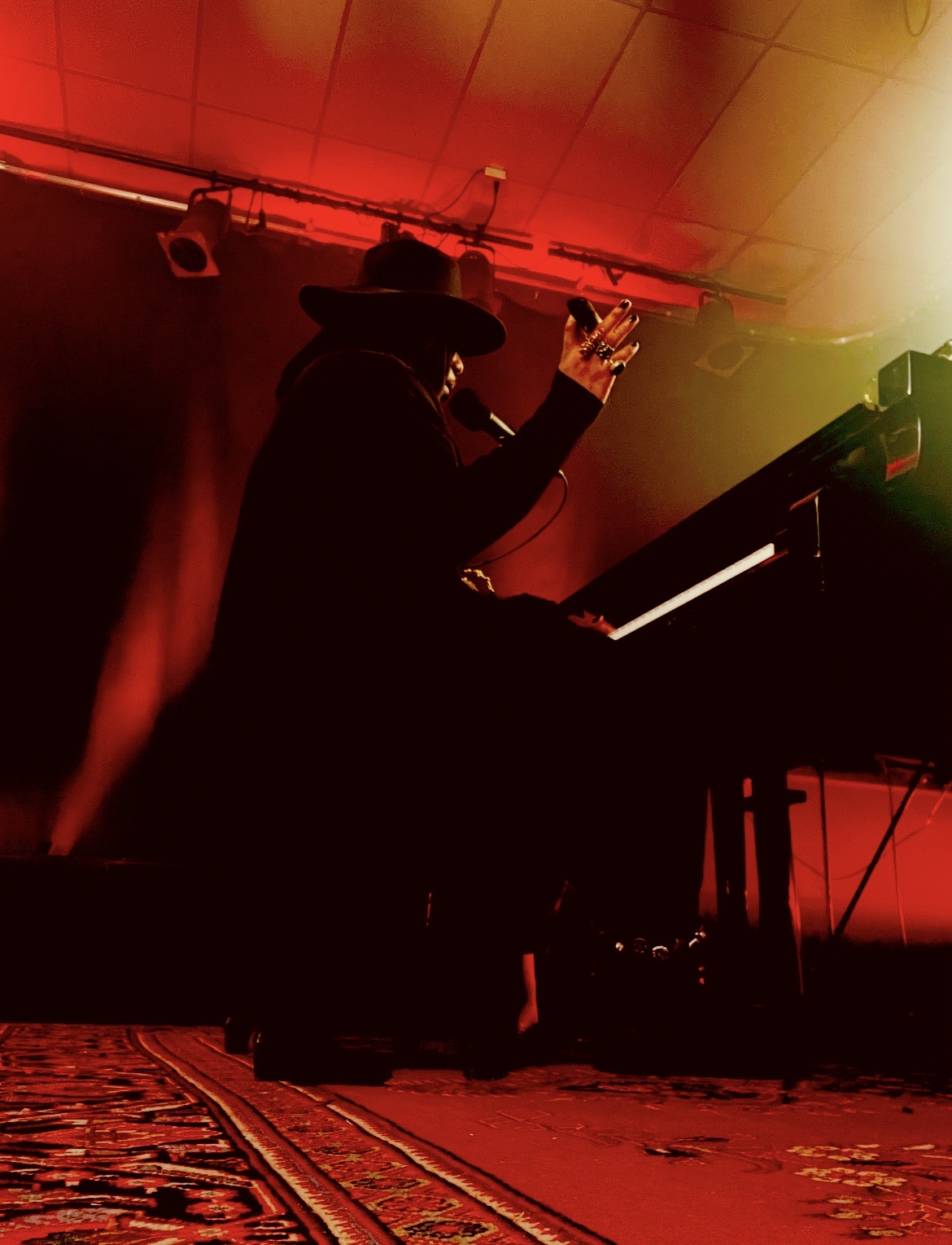 Silhouette of a person playing piano on stage with red and yellow lighting, holding a glass, with a patterned rug visible.