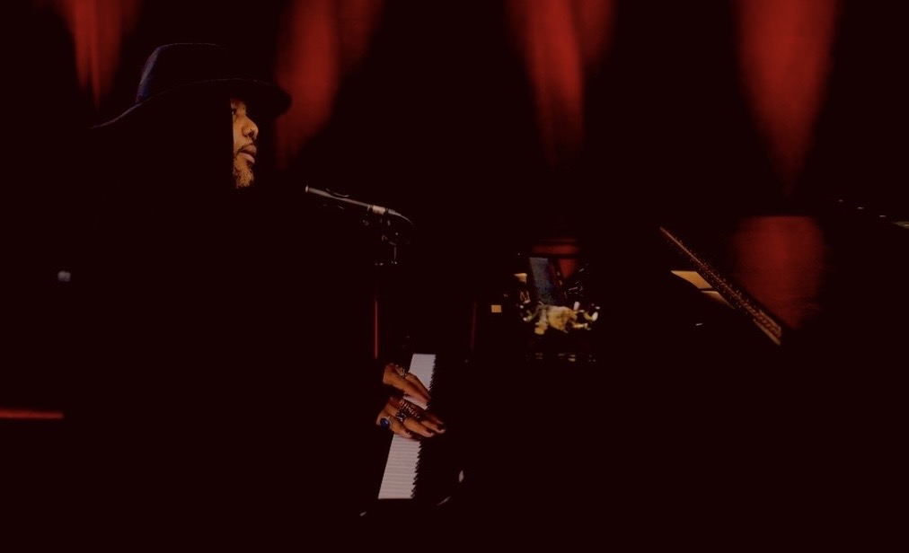 Person playing piano and singing into microphone on stage with red curtains in background.