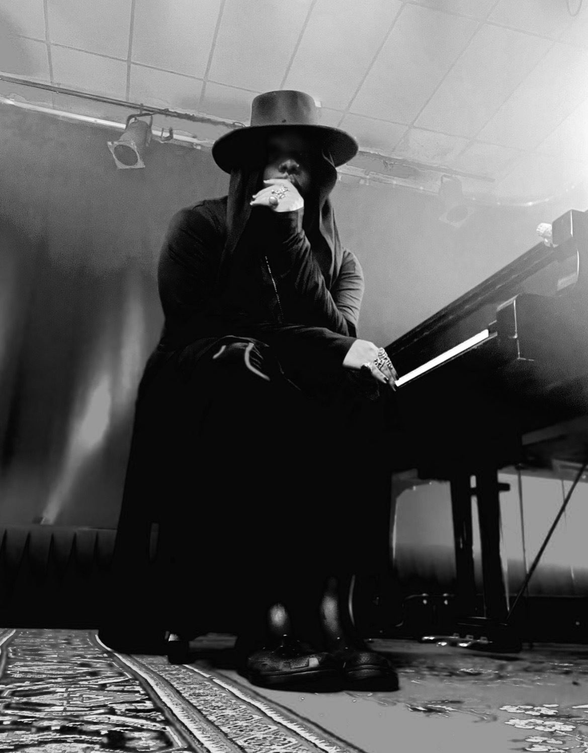 Person wearing a wide-brimmed hat sitting at a piano, holding a microphone, in a dimly lit room