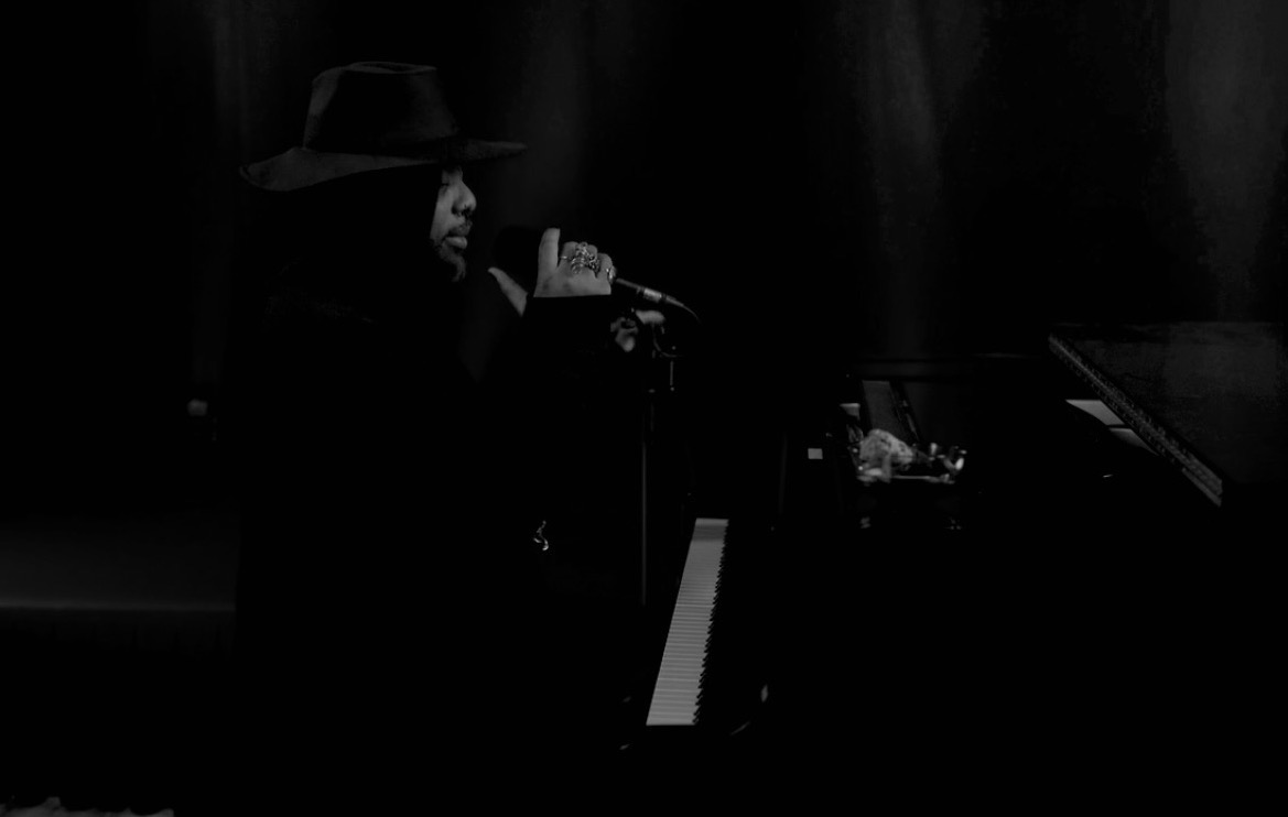 Person wearing a hat singing into a microphone on stage, with a piano nearby, in a dark setting.