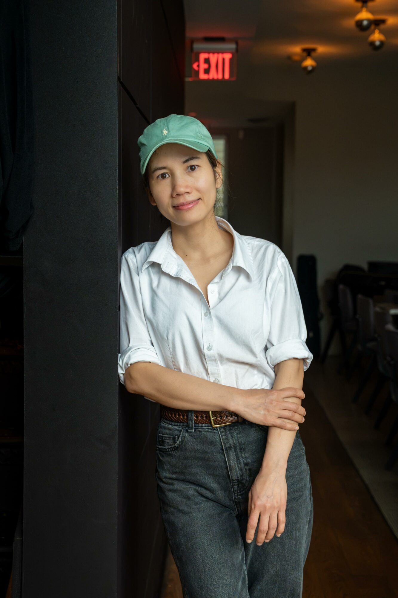 Young woman in white shirt and green cap standing indoors near a black wall, with an exit sign in background.
