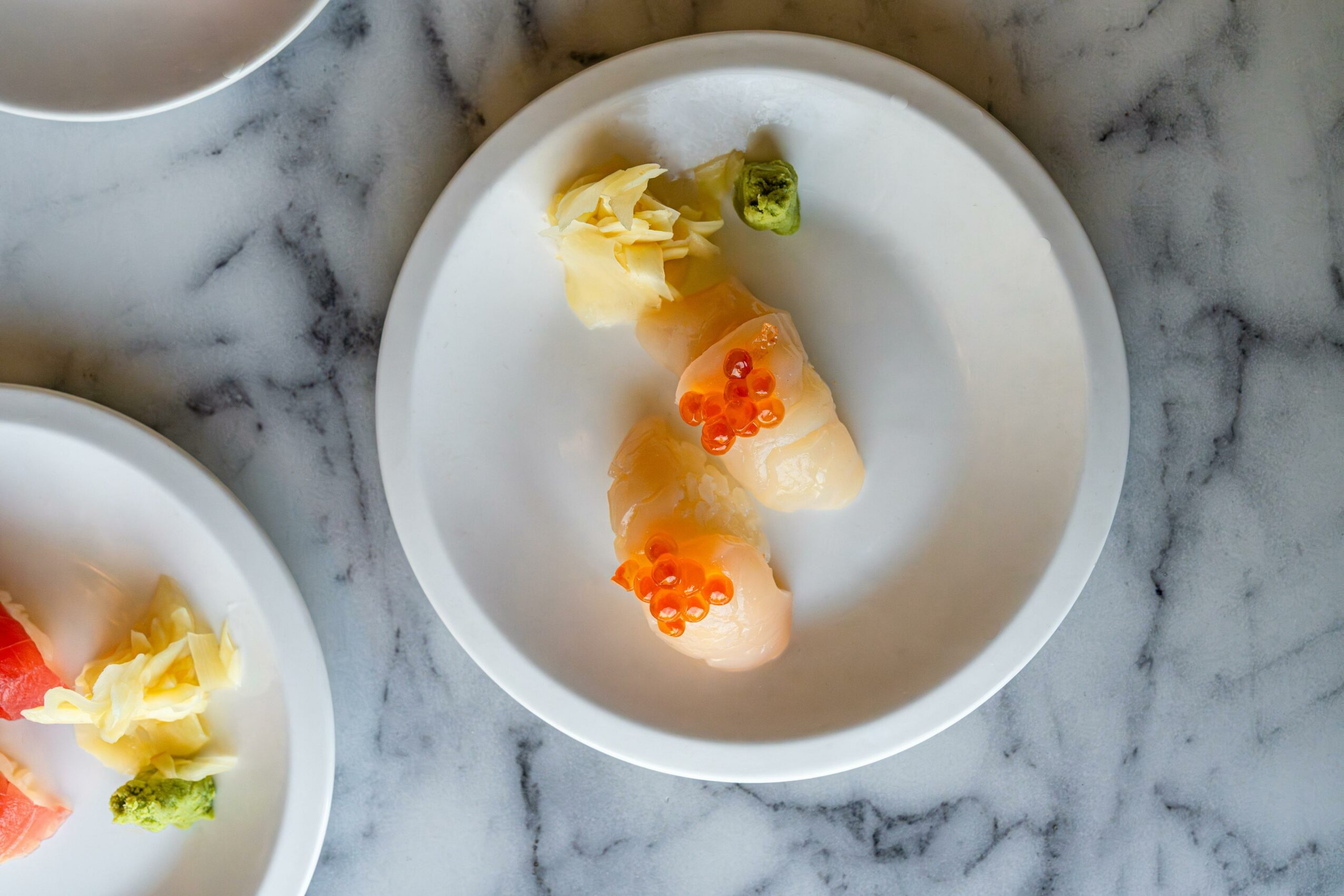 Two pieces of sushi with orange fish roe, garnished with wasabi and ginger on a white plate, marble surface background.