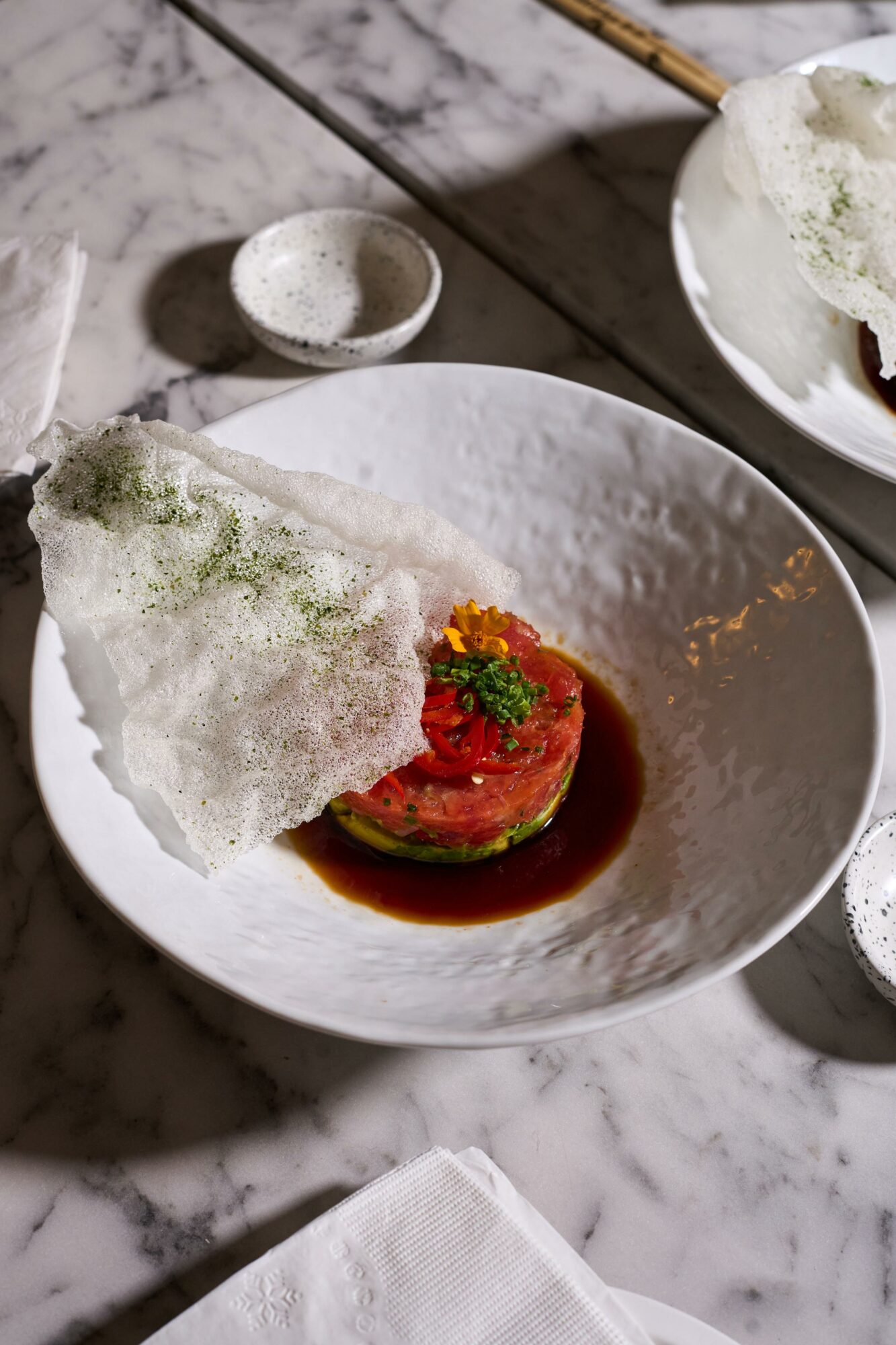 A dish with a round piece of raw meat topped with herbs, served in a white bowl with sauce, and a crispy garnish.