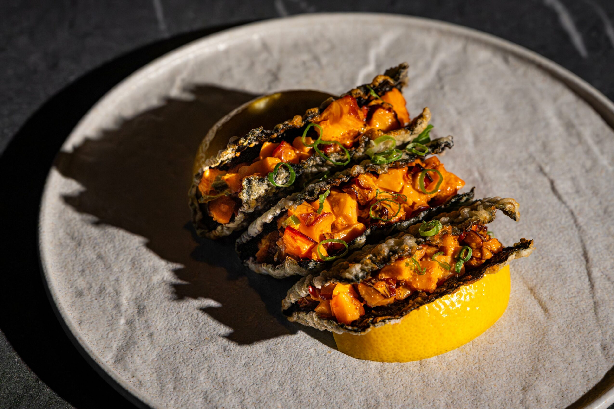 Three pieces of sushi with orange filling and green topping on a white plate, with a lemon wedge underneath, in a dark setting.