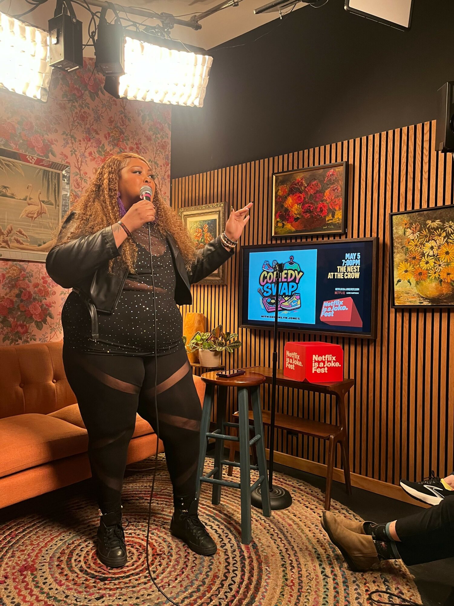 Woman with long hair in black outfit standing in front of a TV screen, talking on a phone, in a decorated room.