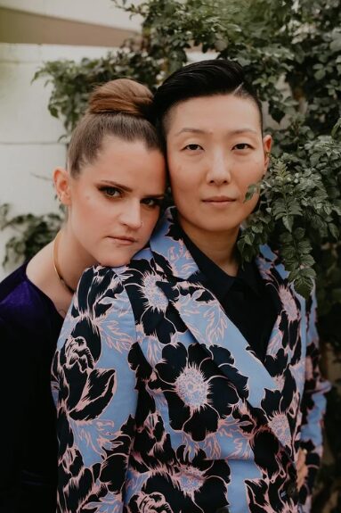 Two women standing close together outdoors, one with dark hair and the other with brown hair in a bun, surrounded by greenery.
