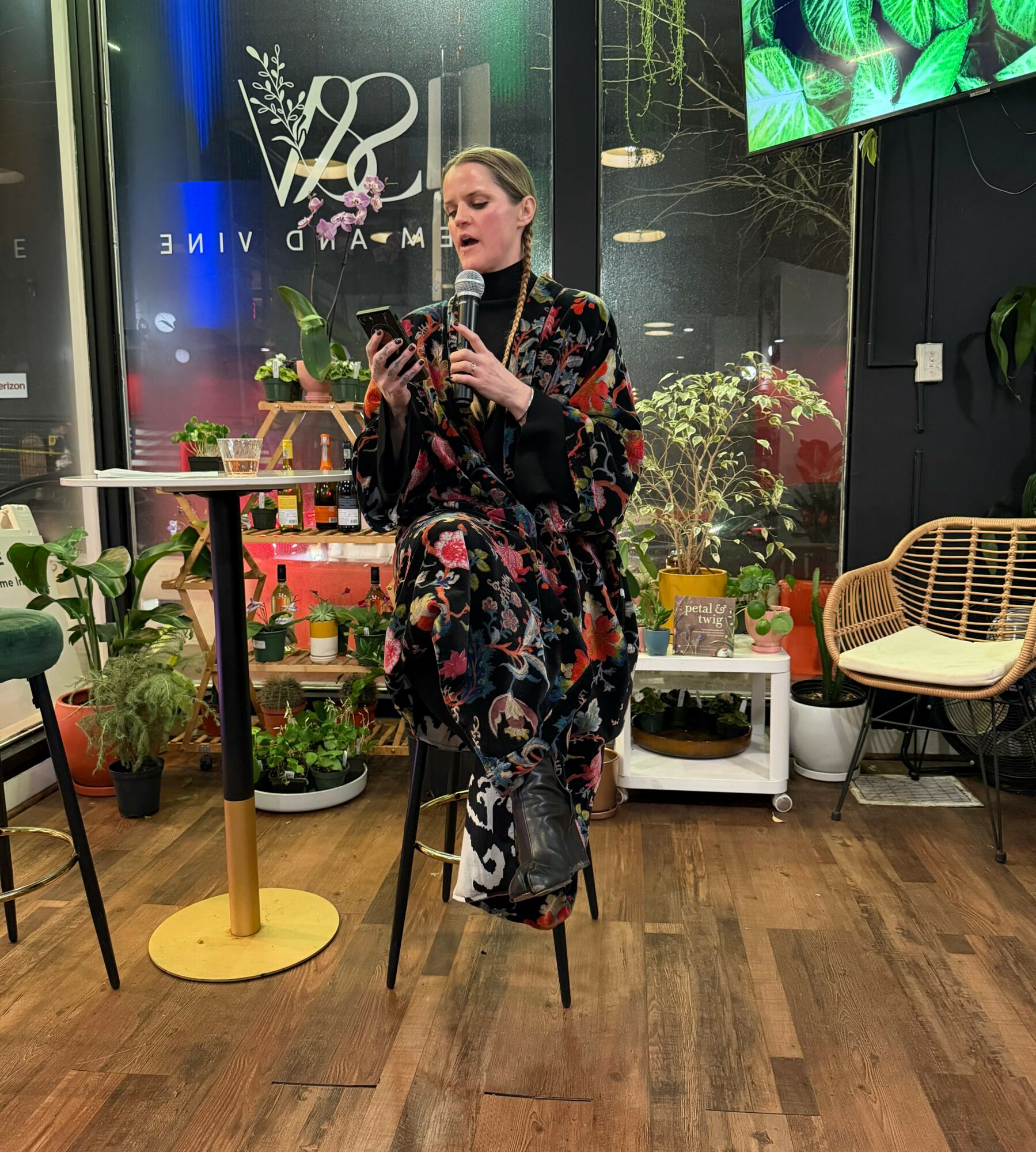 Woman sitting on a stool, reading from a phone in a cozy indoor space with plants and a chair.