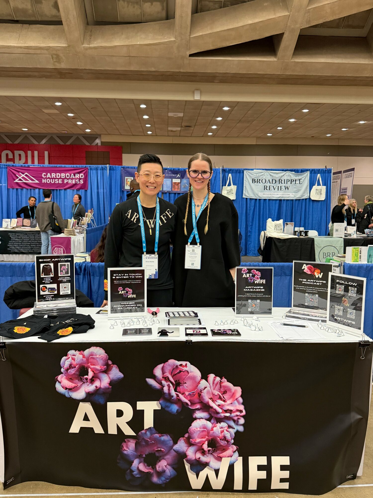 Two women standing behind a table with art and wedding information at an indoor event, smiling, with booths in the background.