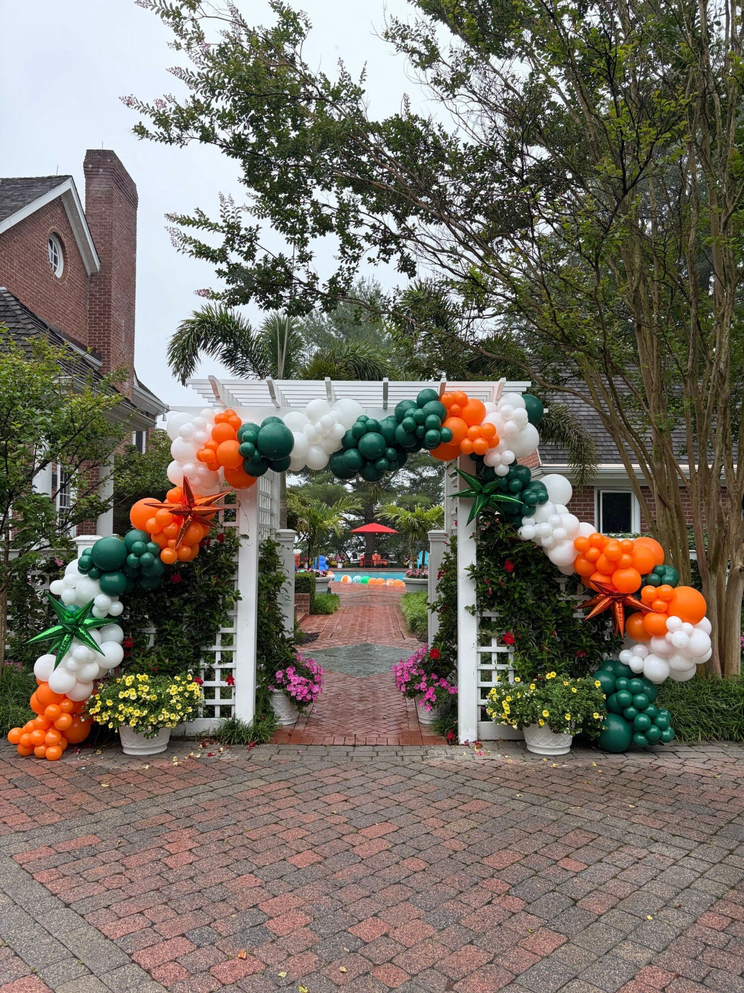 Decorated archway with balloons in orange, white, green, and teal, surrounded by trees and flowers, on a brick pathway.