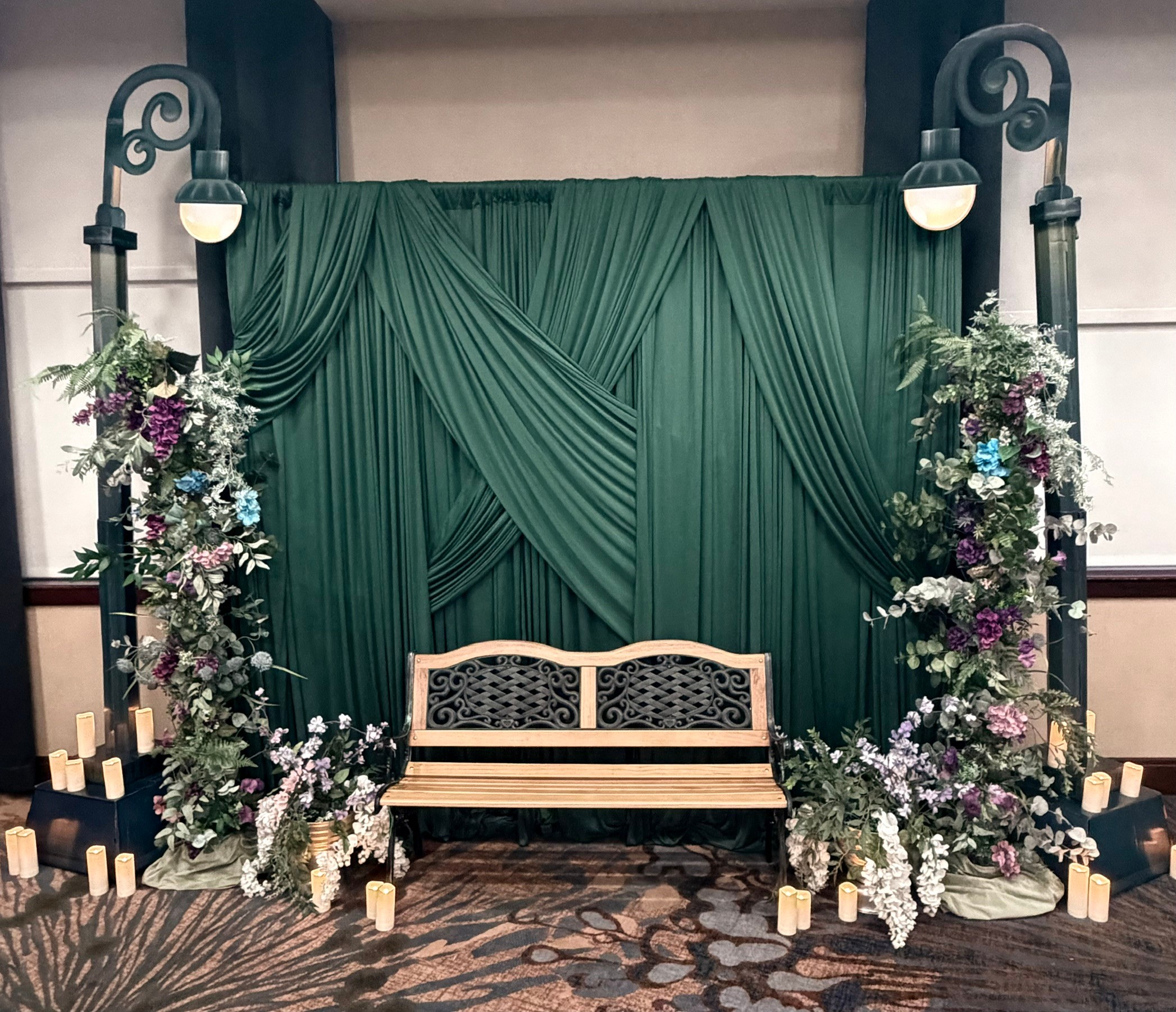 Green draped backdrop with floral arrangements and a wooden bench in front, candles on the floor, in an indoor setting.