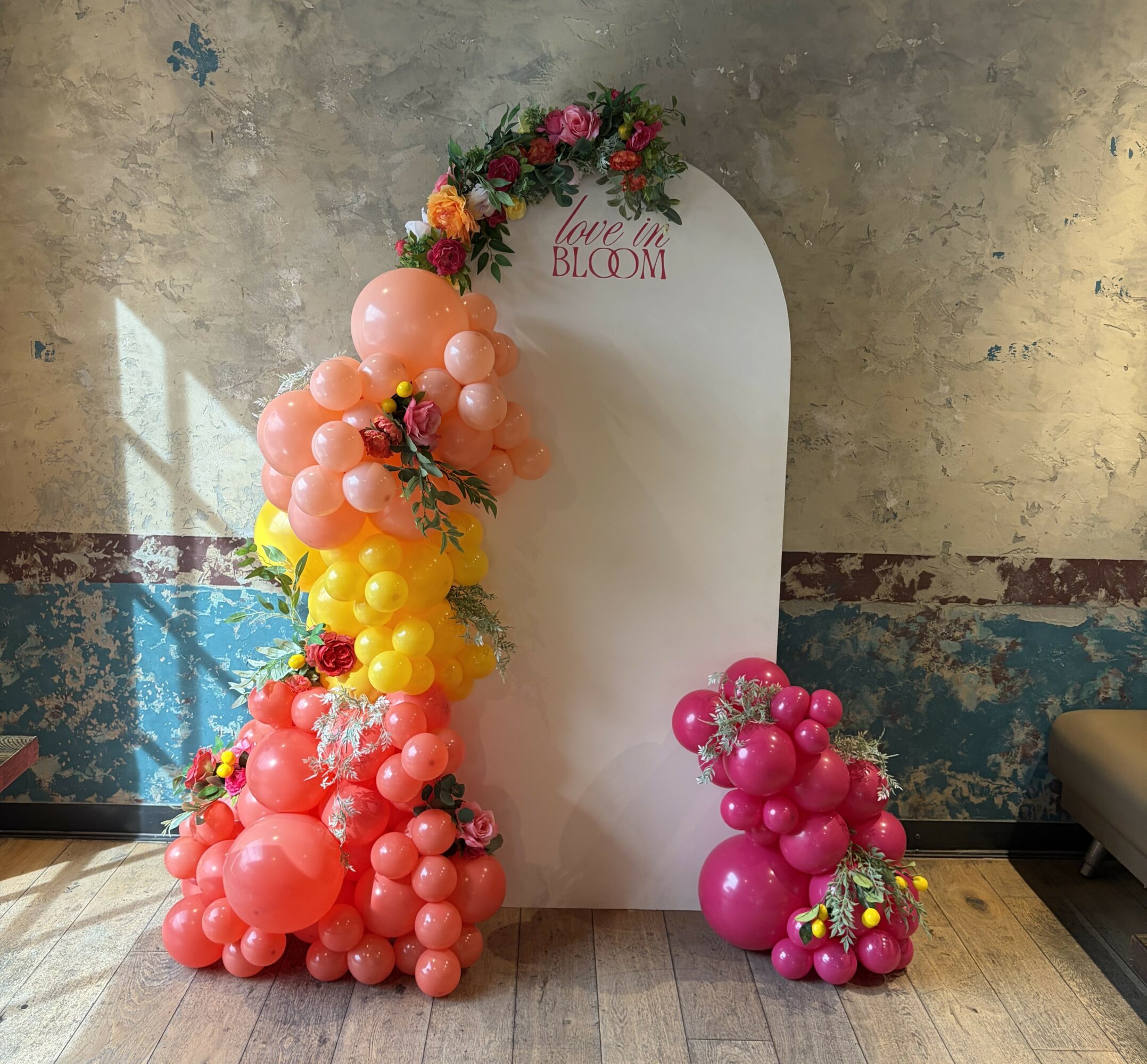Balloon arrangements with pink, yellow, and orange balloons, flowers, and a white backdrop with text, on a wooden floor.