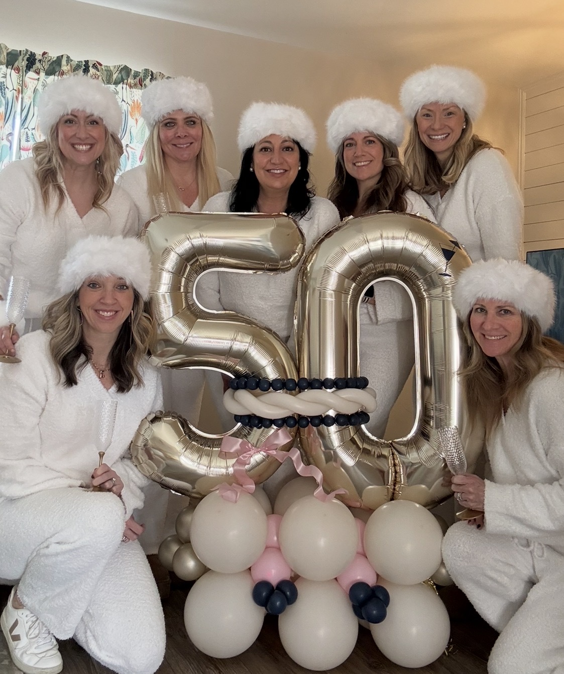 Group of seven women in white outfits and fluffy hats celebrating with balloons and large silver '50' numbers.