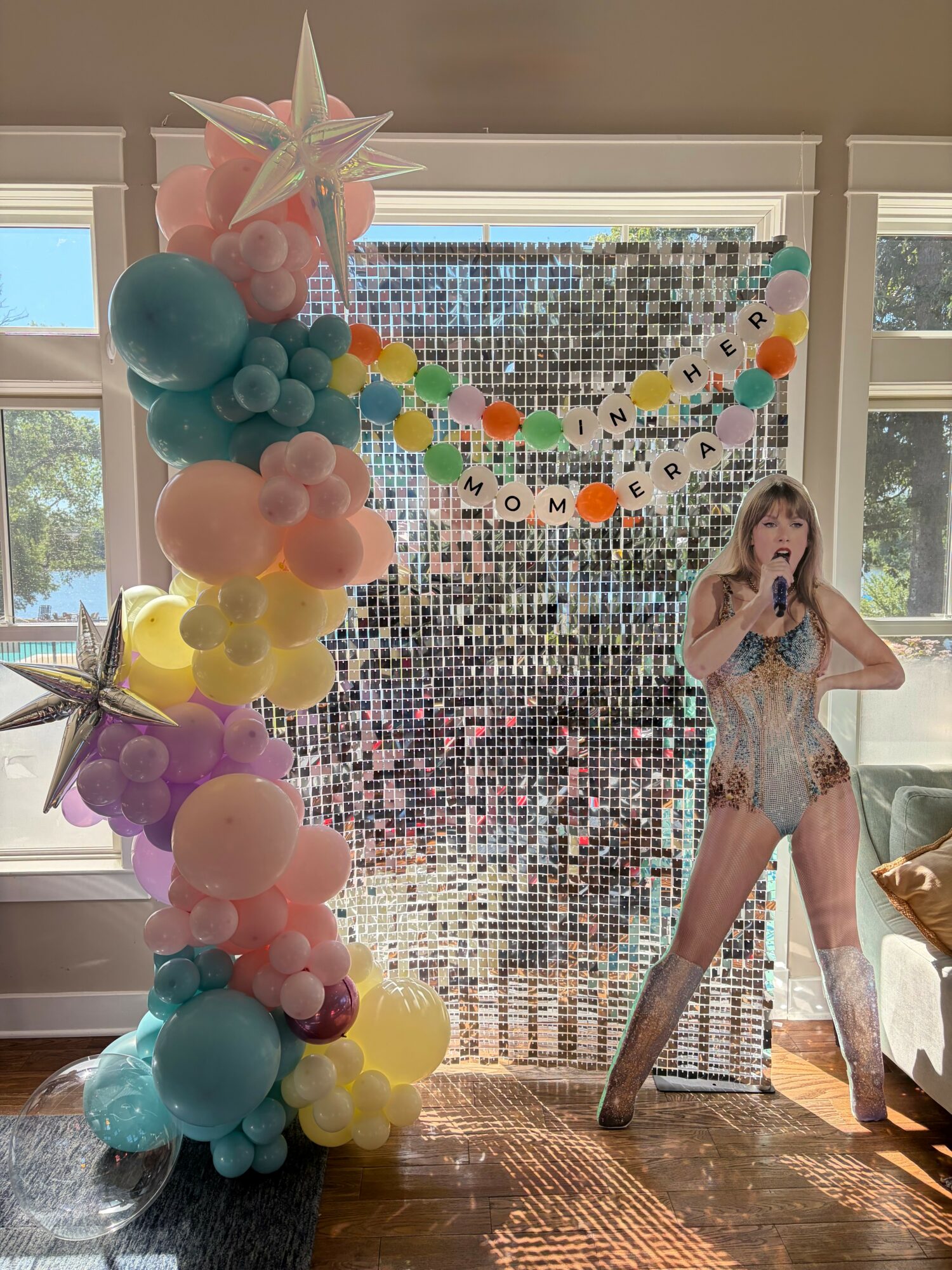 Person in a sparkly outfit standing next to a colorful balloon arrangement and a shiny backdrop, holding a microphone.
