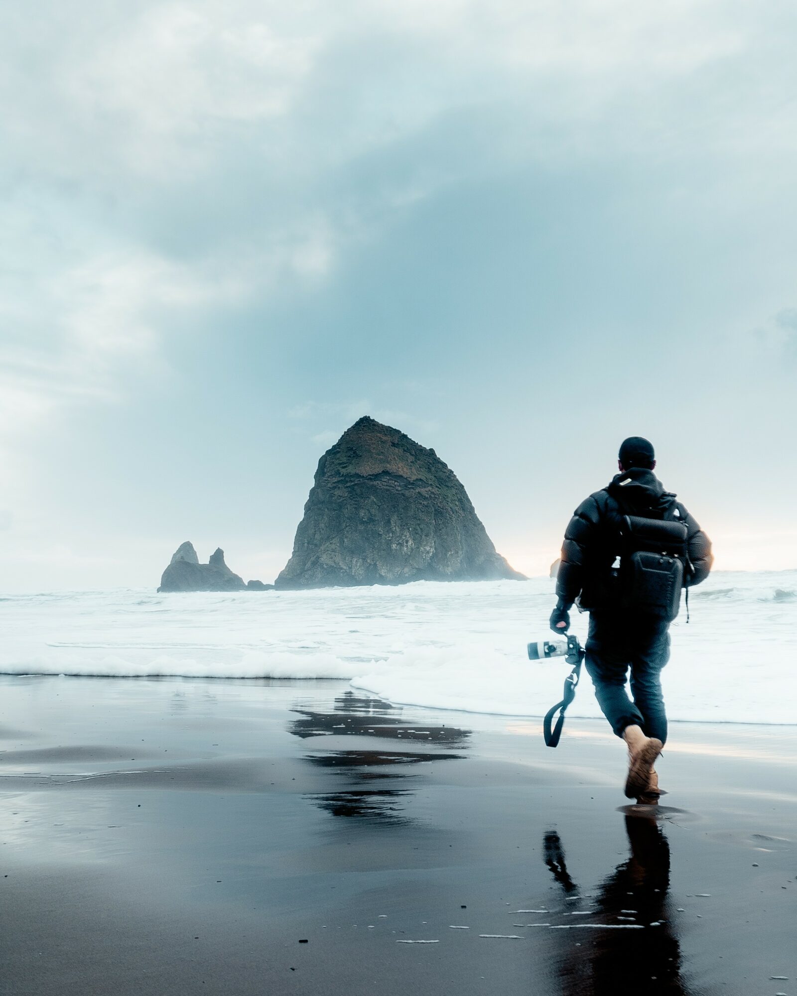 Person walking on wet beach with large rock formation in ocean background, cloudy sky, reflection on wet sand.