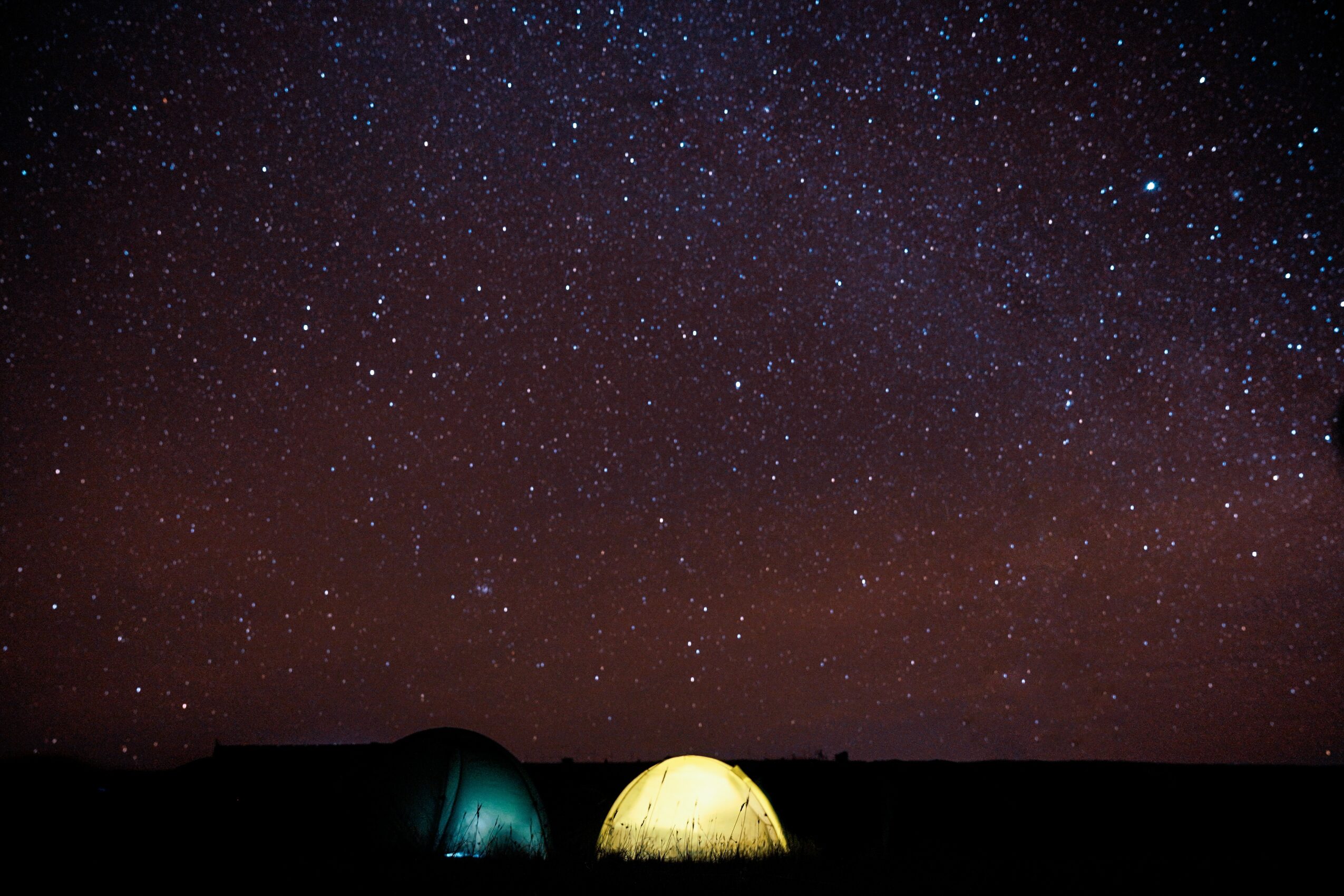 Night sky filled with stars above two tents on a dark landscape, one tent glowing yellow and the other teal.
