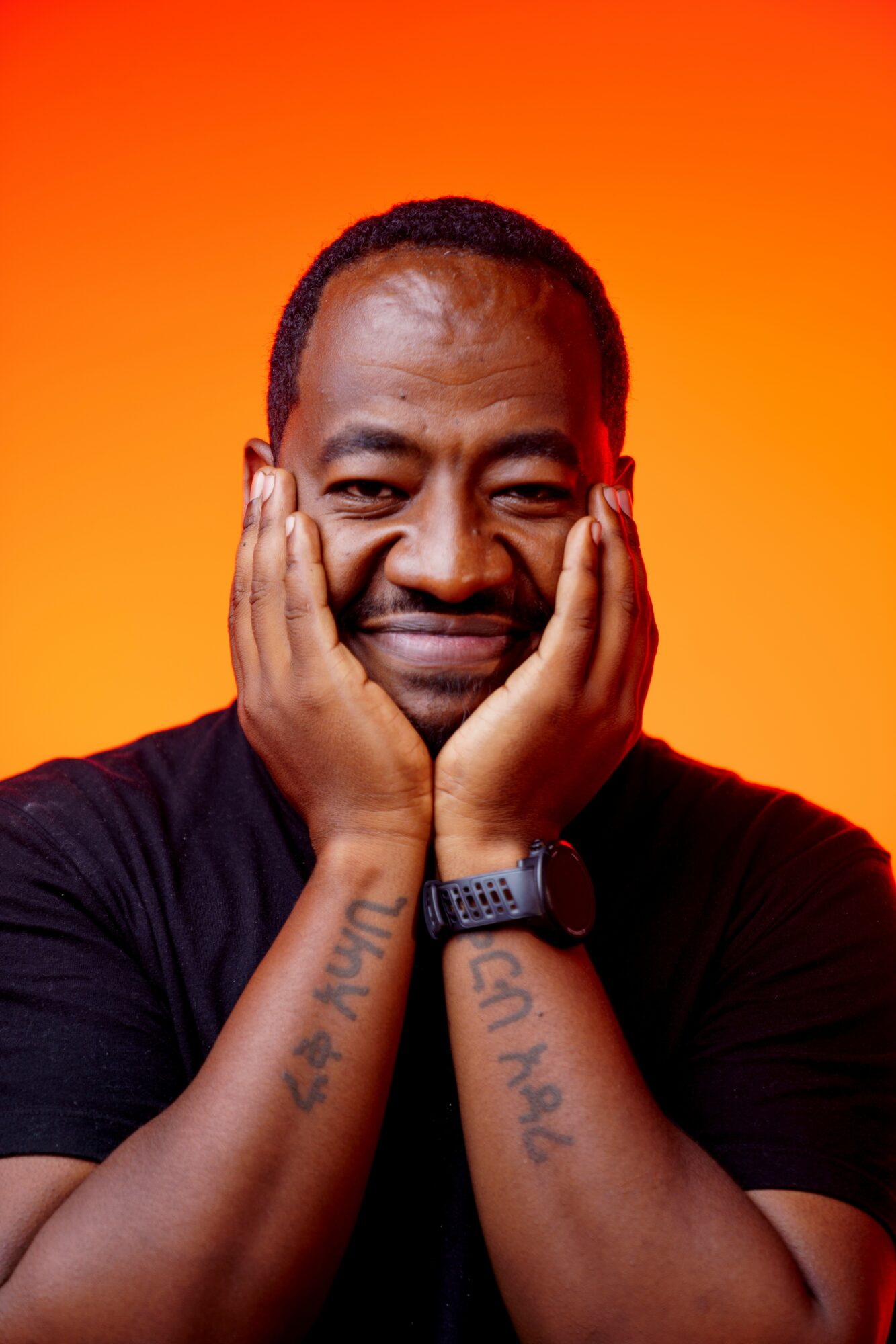 Person with hands on face, smiling, against orange background, wearing a black shirt and watch, with tattoos on arms.