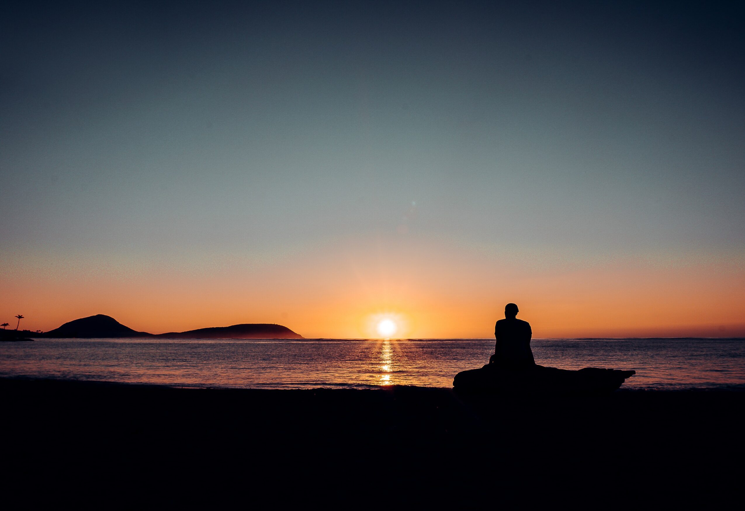 Person sitting on a rock watching a sunset over the ocean with distant islands, silhouetted against the sky.