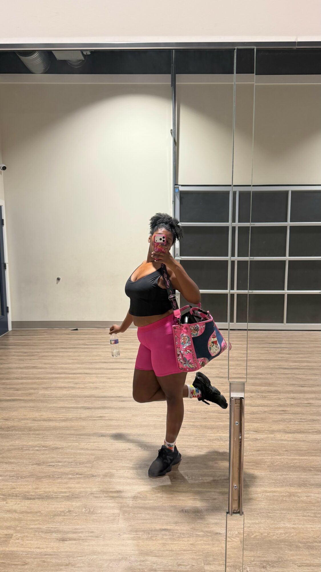 Person taking a mirror selfie in a room with wooden floor, black and white wall tiles, holding a pink bag, wearing black top and pink shorts.