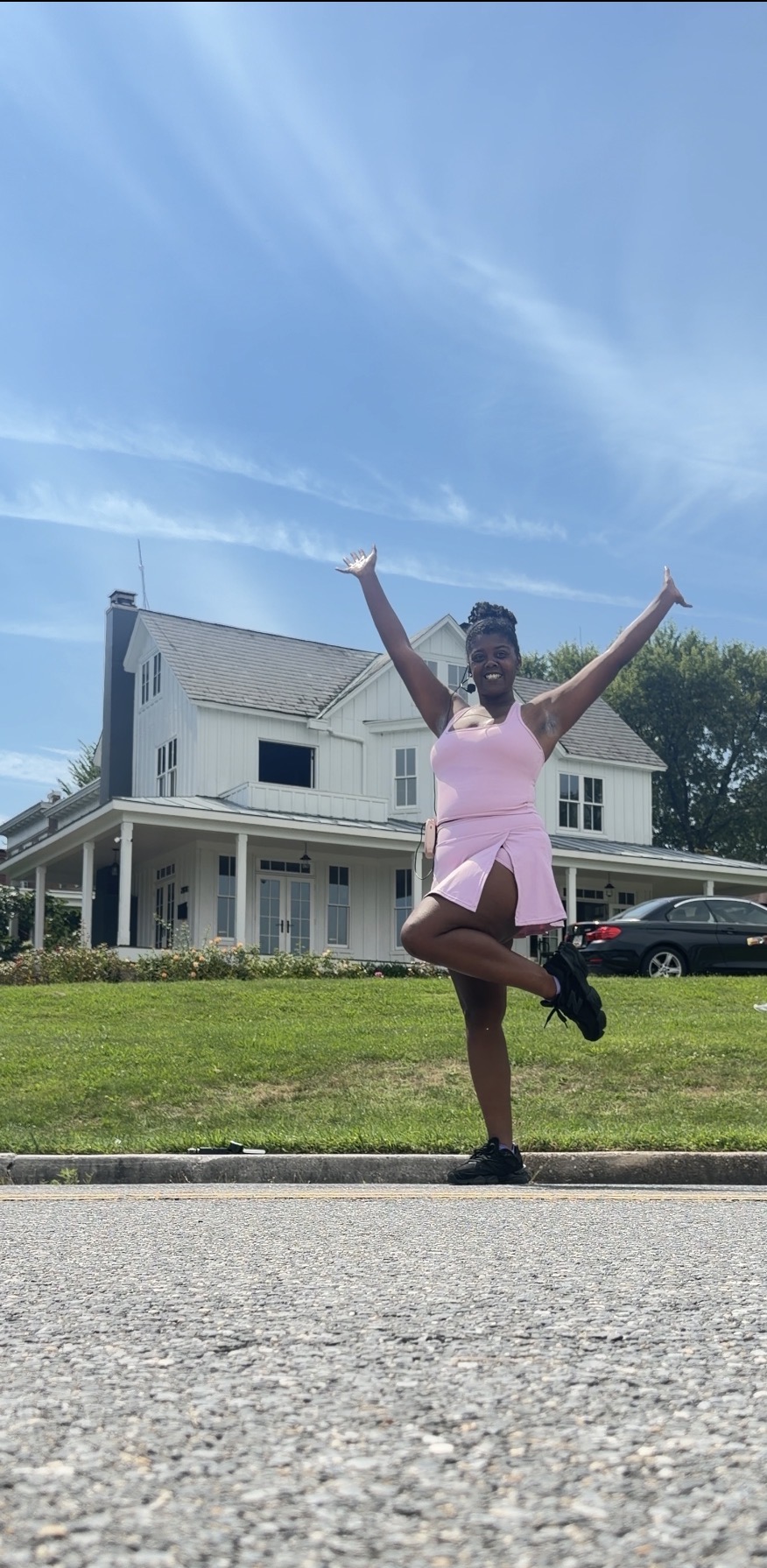 Person in pink dress performing a yoga pose outdoors in front of a house, with arms raised and one leg bent.