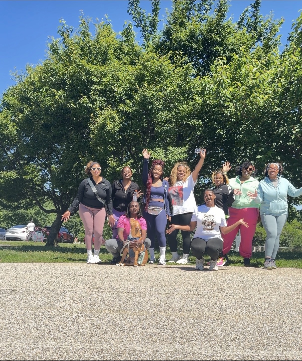 Group of nine women and children outdoors on grass with trees and blue sky, some smiling and waving.