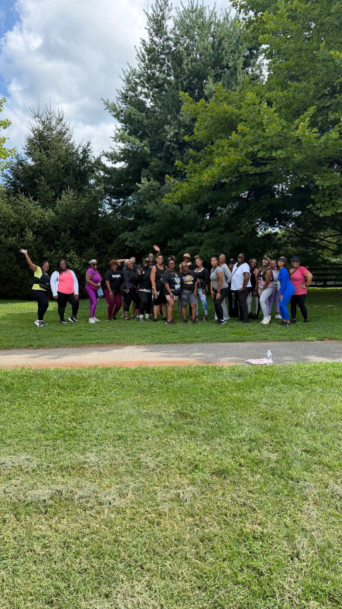 Group of people standing outdoors under trees on grass, some holding objects, with cloudy sky above.