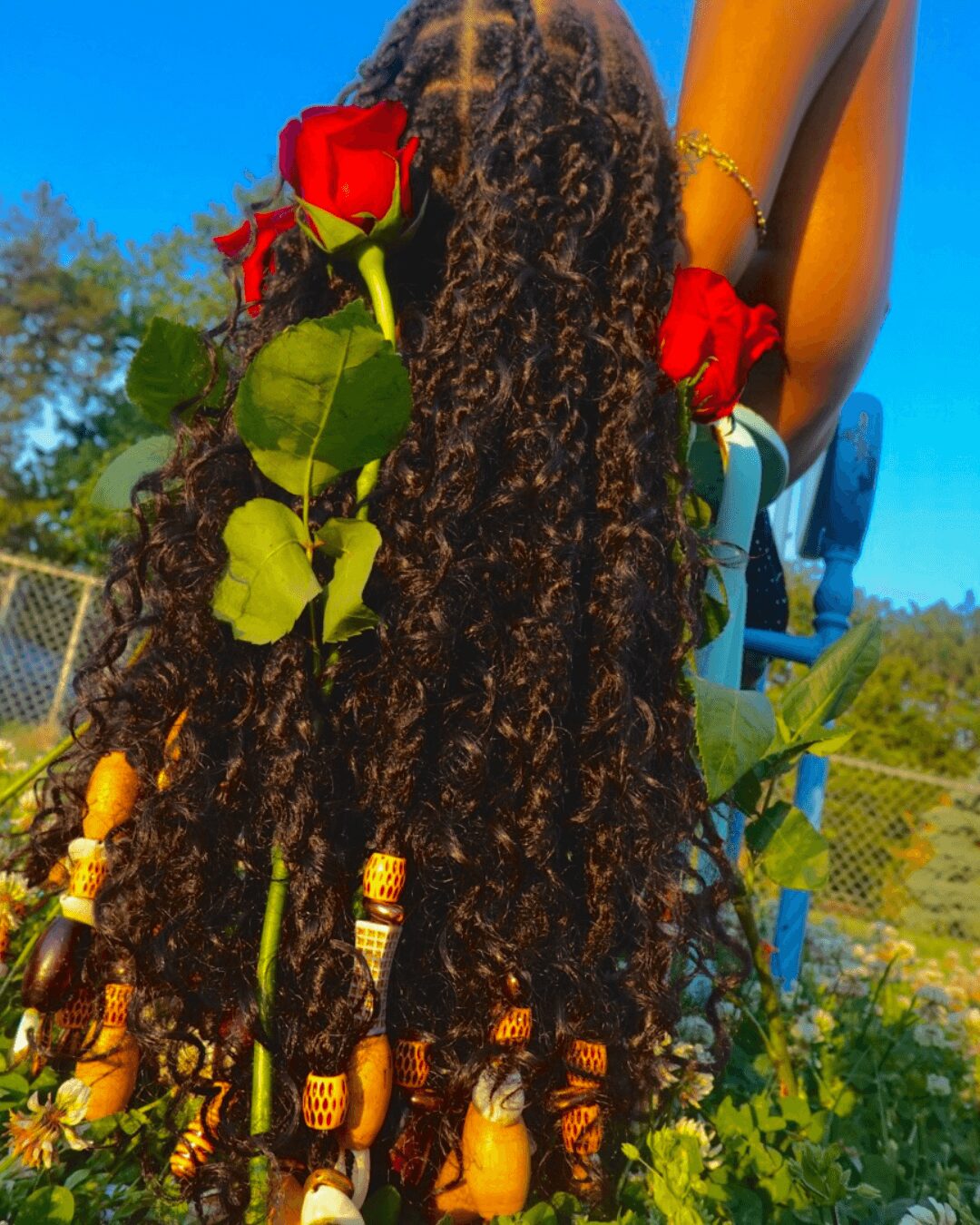 Person with long, curly hair holding red roses outdoors against a blue sky and greenery.