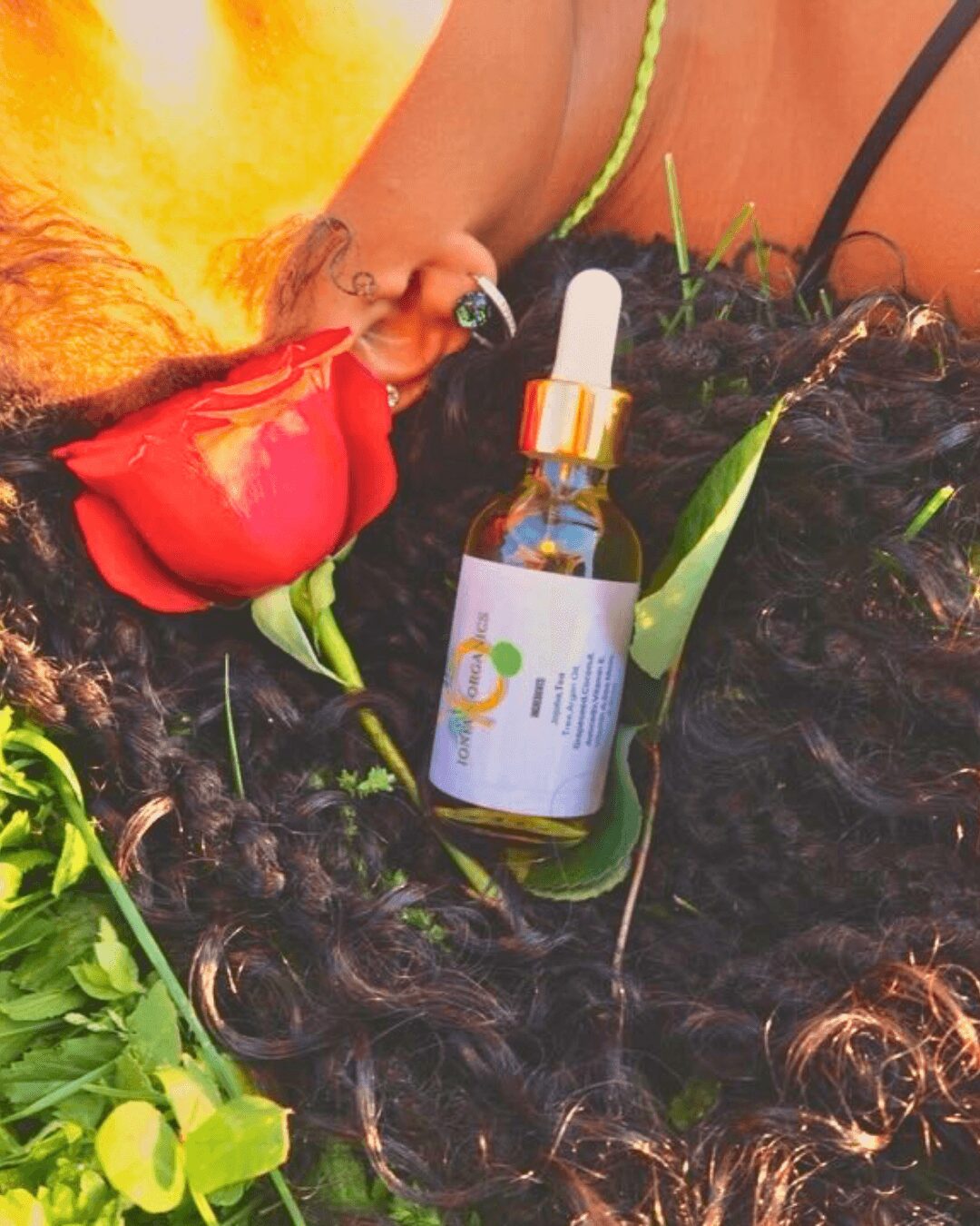 A small glass dropper bottle with a white label and orange cap placed on dark curly hair surrounded by green leaves and a red flower petal.