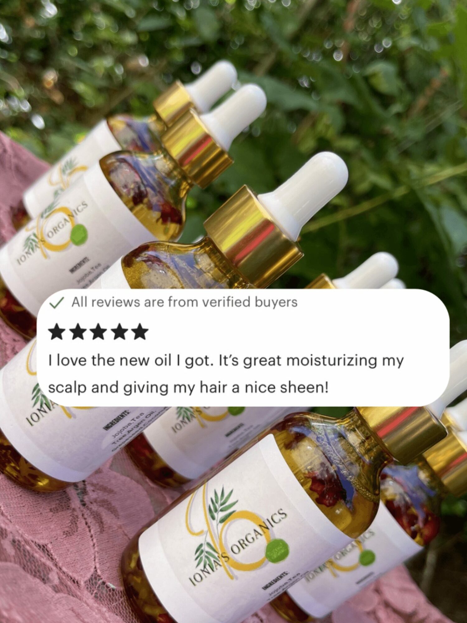 Four bottles of hair oil with white labels and gold caps placed outdoors on a pink cloth, surrounded by green foliage.
