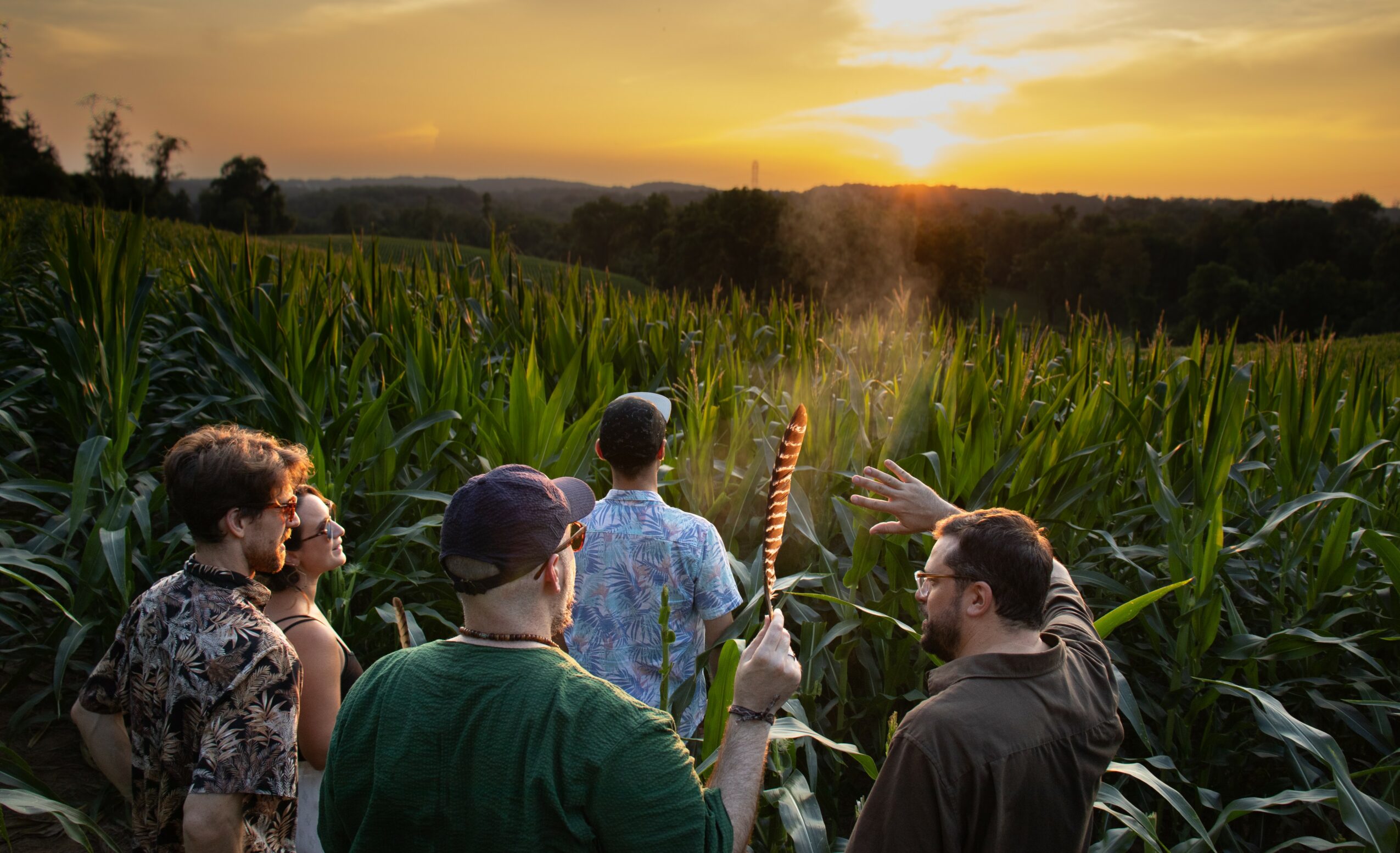 Group of people standing in a cornfield during sunset, some holding a torch, with a landscape in the background.