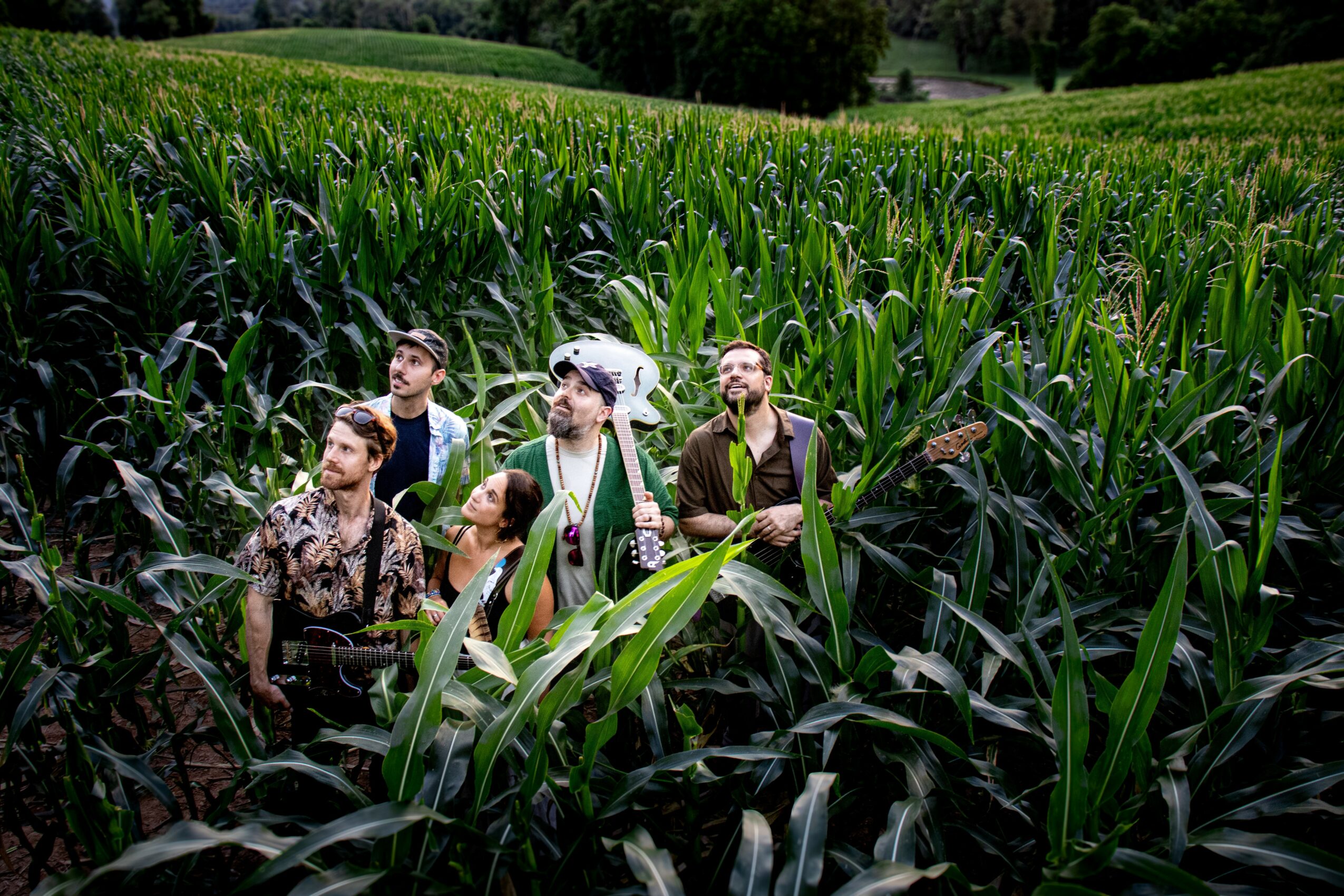 Group of five people standing in a tall green cornfield with trees and hills in the background.