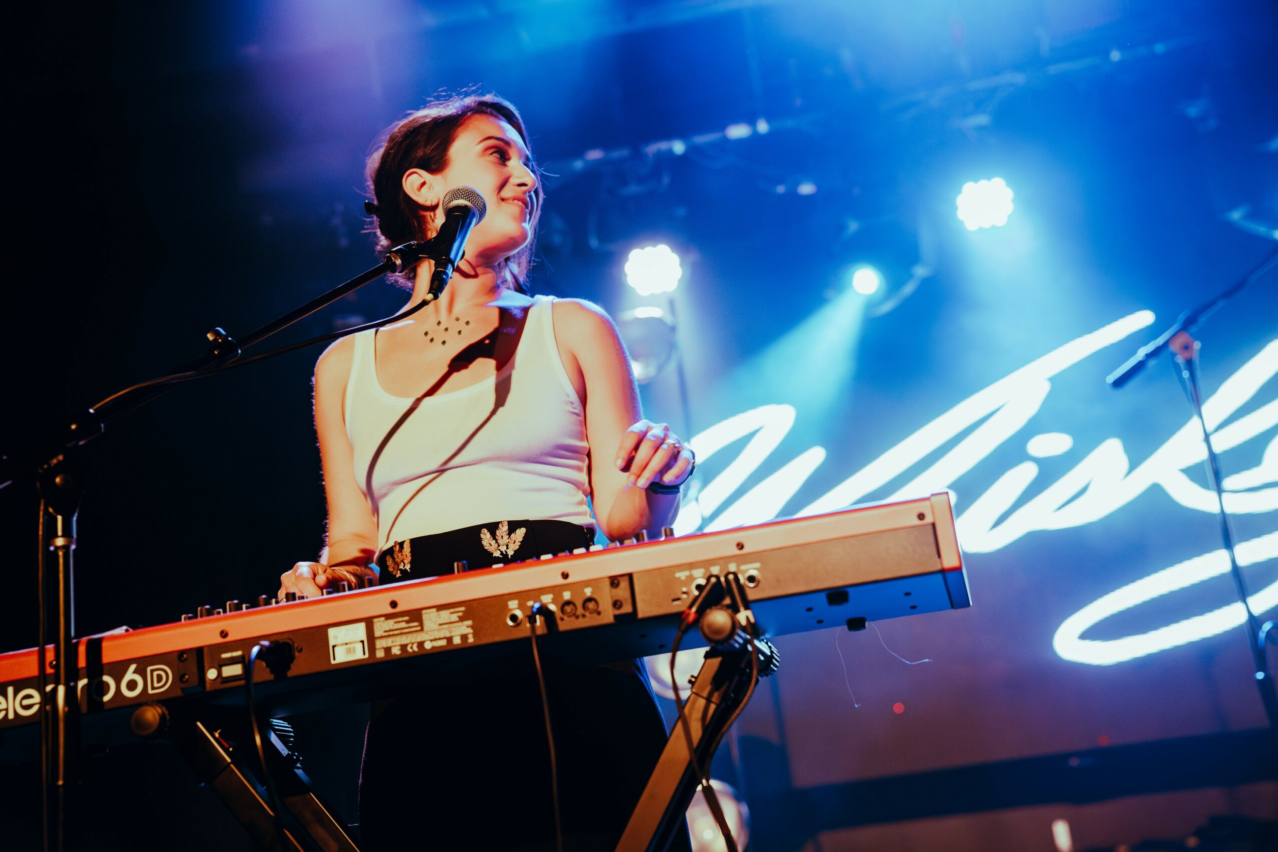 A woman plays a keyboard on stage with blue and purple lighting, smiling and looking to the side.