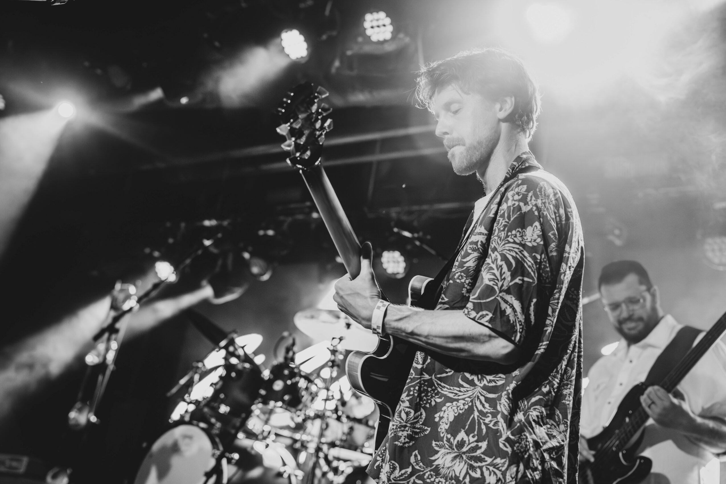 Person playing guitar on stage with bright lights and a band in the background, black and white photo
