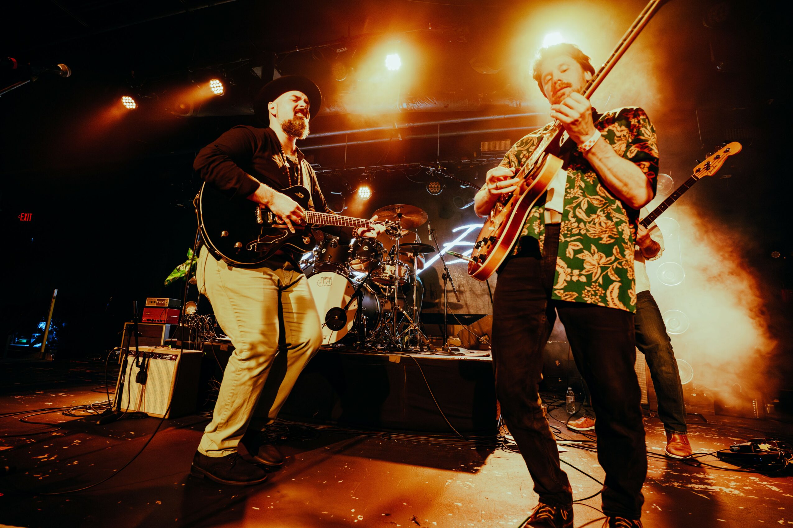 Two musicians playing guitars on stage with bright lights and smoke effects, one wearing a floral shirt and the other in dark clothing.