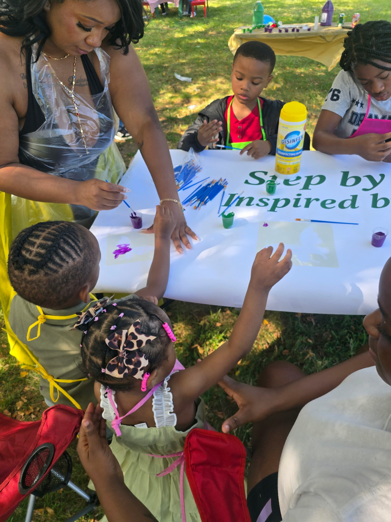 Children and an adult painting on a large white banner outdoors, with trees and grass in the background.