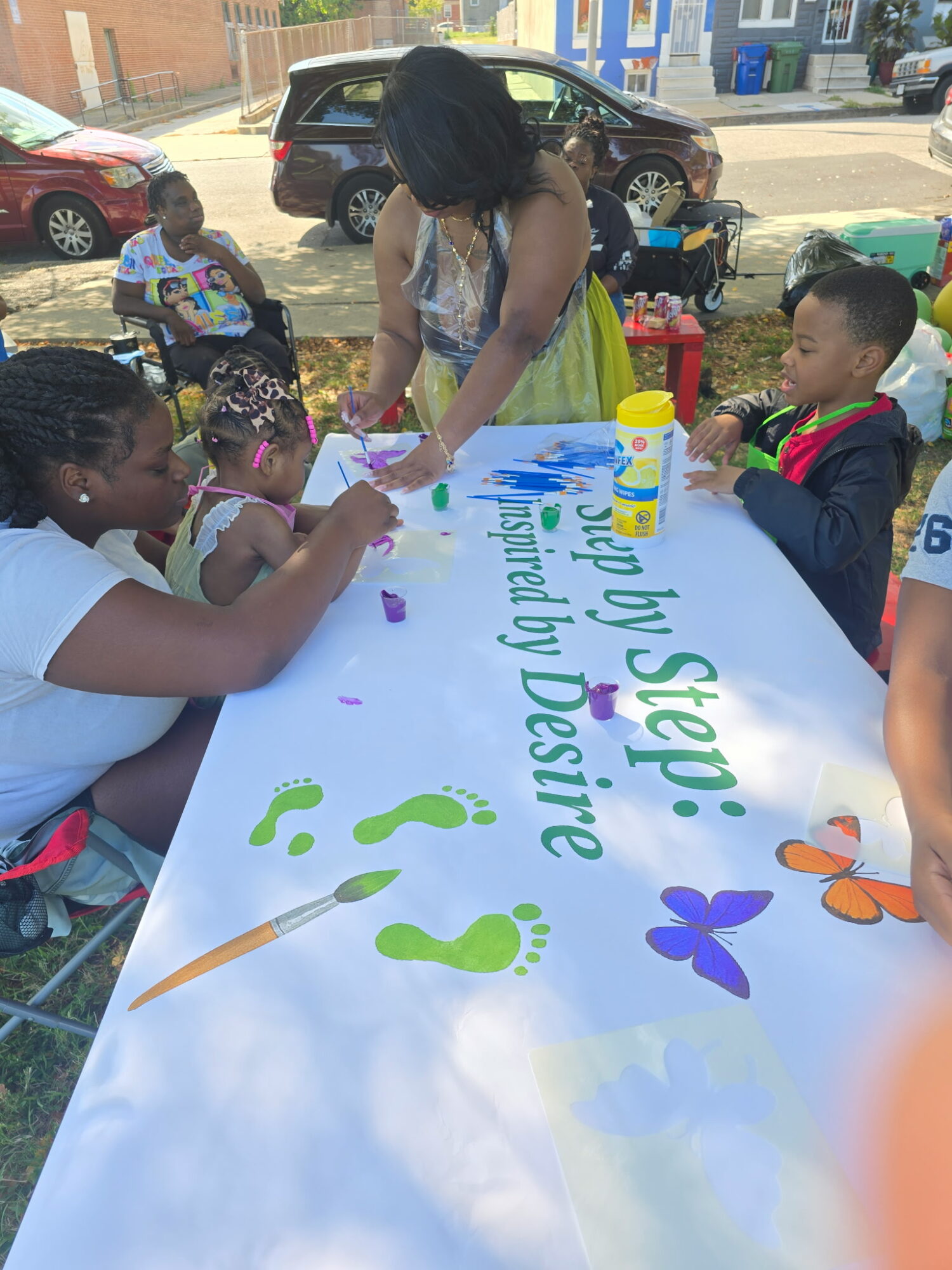 Children and an adult painting a large banner outdoors with colorful text and decorations.